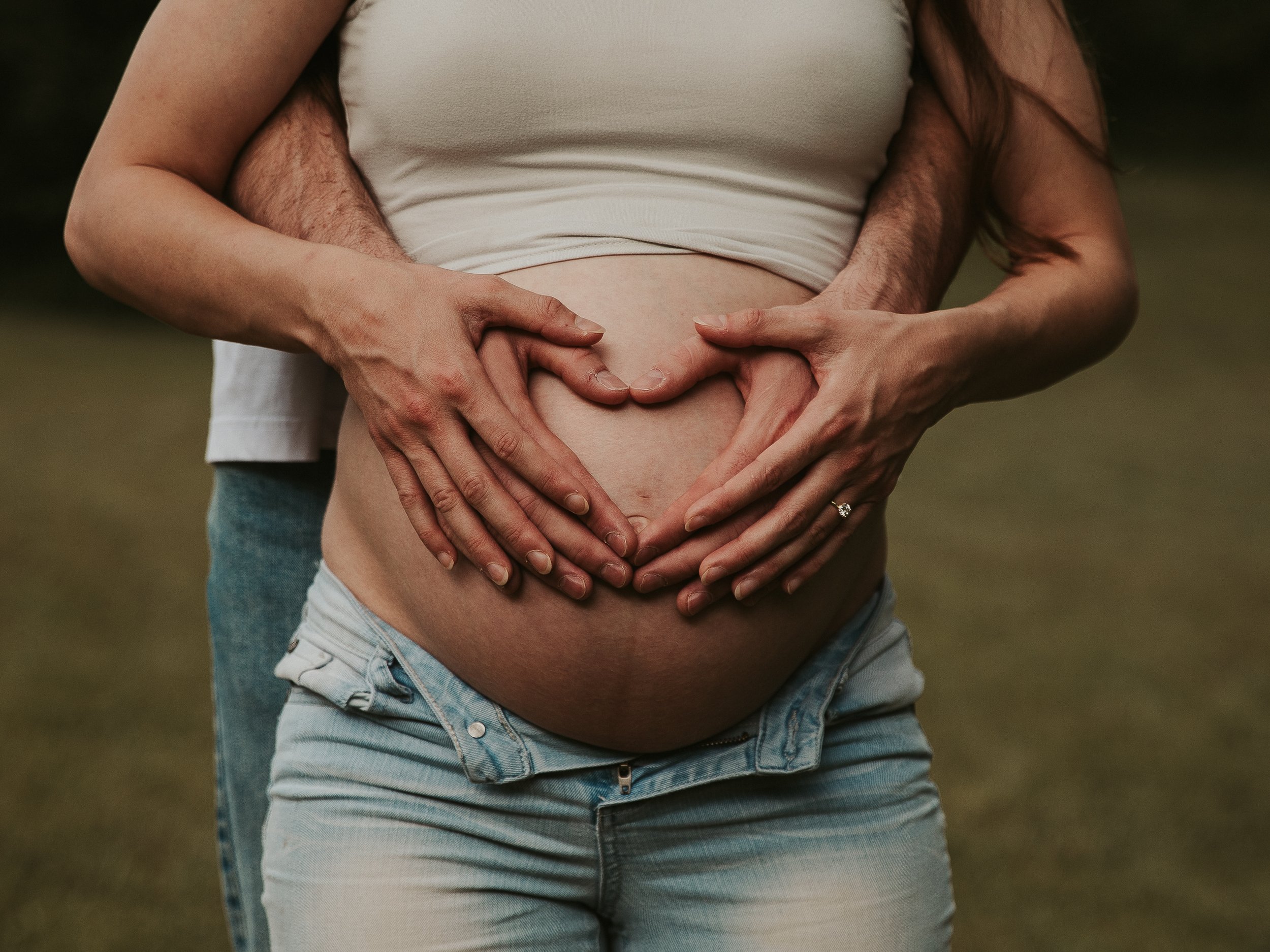 A pregnant woman wearing a white tank top and light blue jeans with the zipper open. A man, partially visible, is standing behind her, holding her belly with both hands. Their hands have cooperation to form a heart shape on her baby bump.