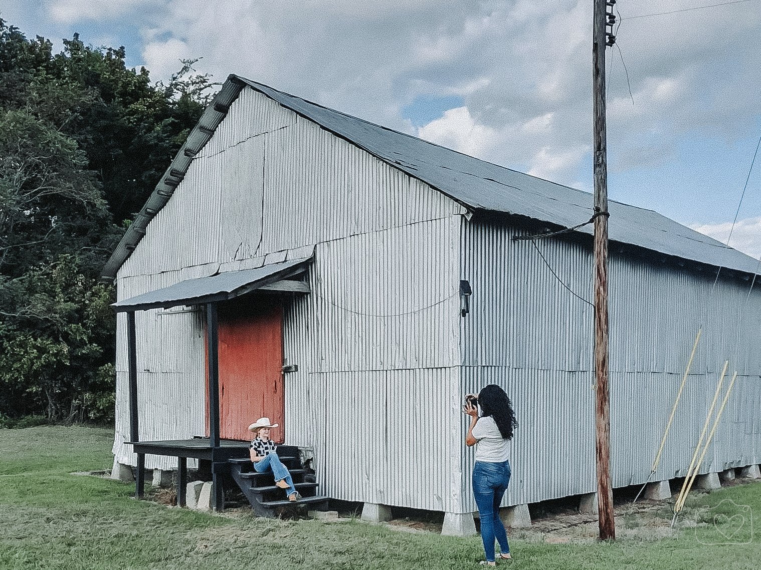 A woman is taking a photo of a young girl sitting on the steps of a small, rustic metal building with a red door. The building is set on a grassy area with trees in the background and an overcast sky overhead.