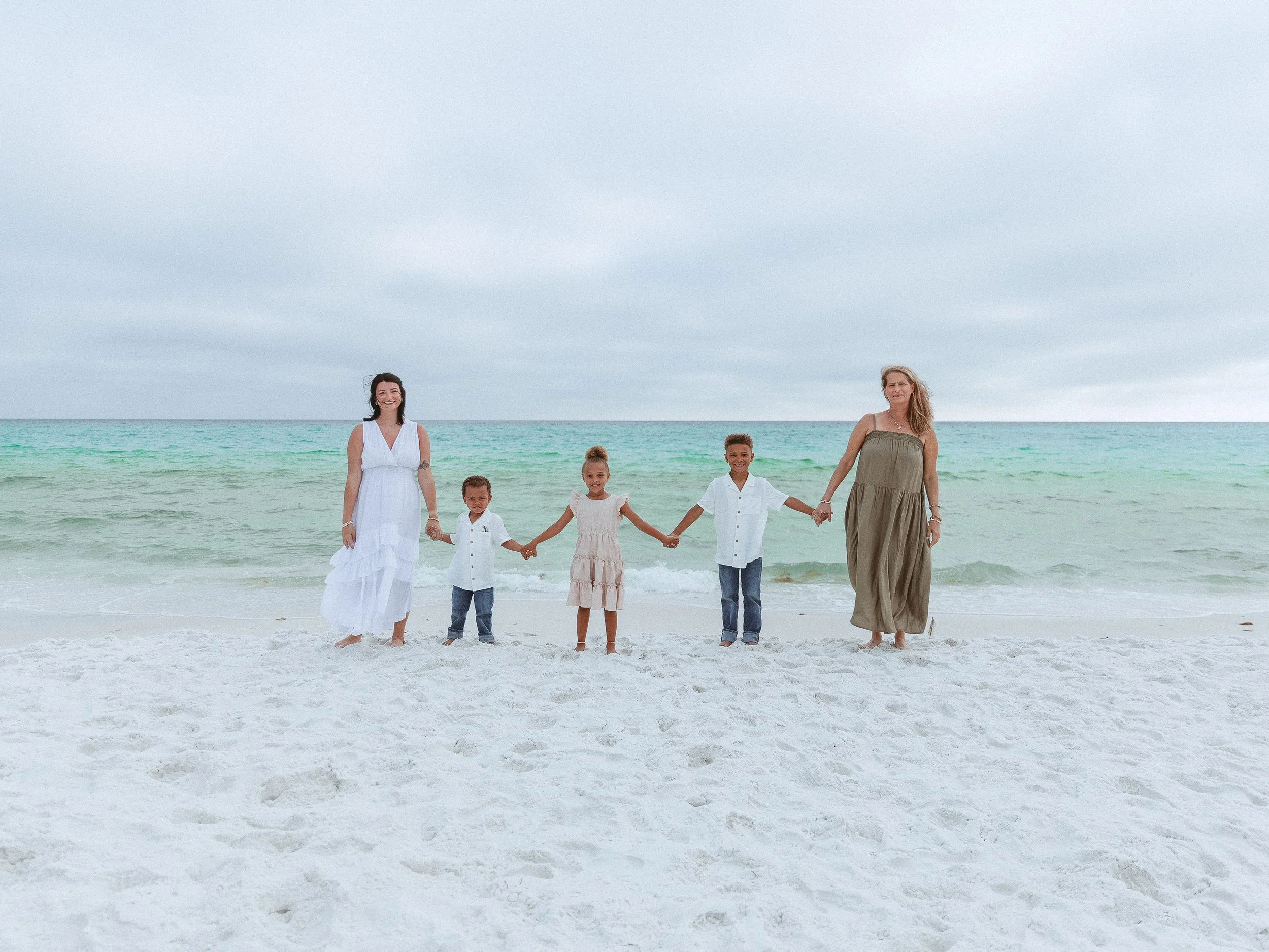 A group of six people, two adults and four children, holding hands and walking along the beach with ocean and cloudy sky in the background.