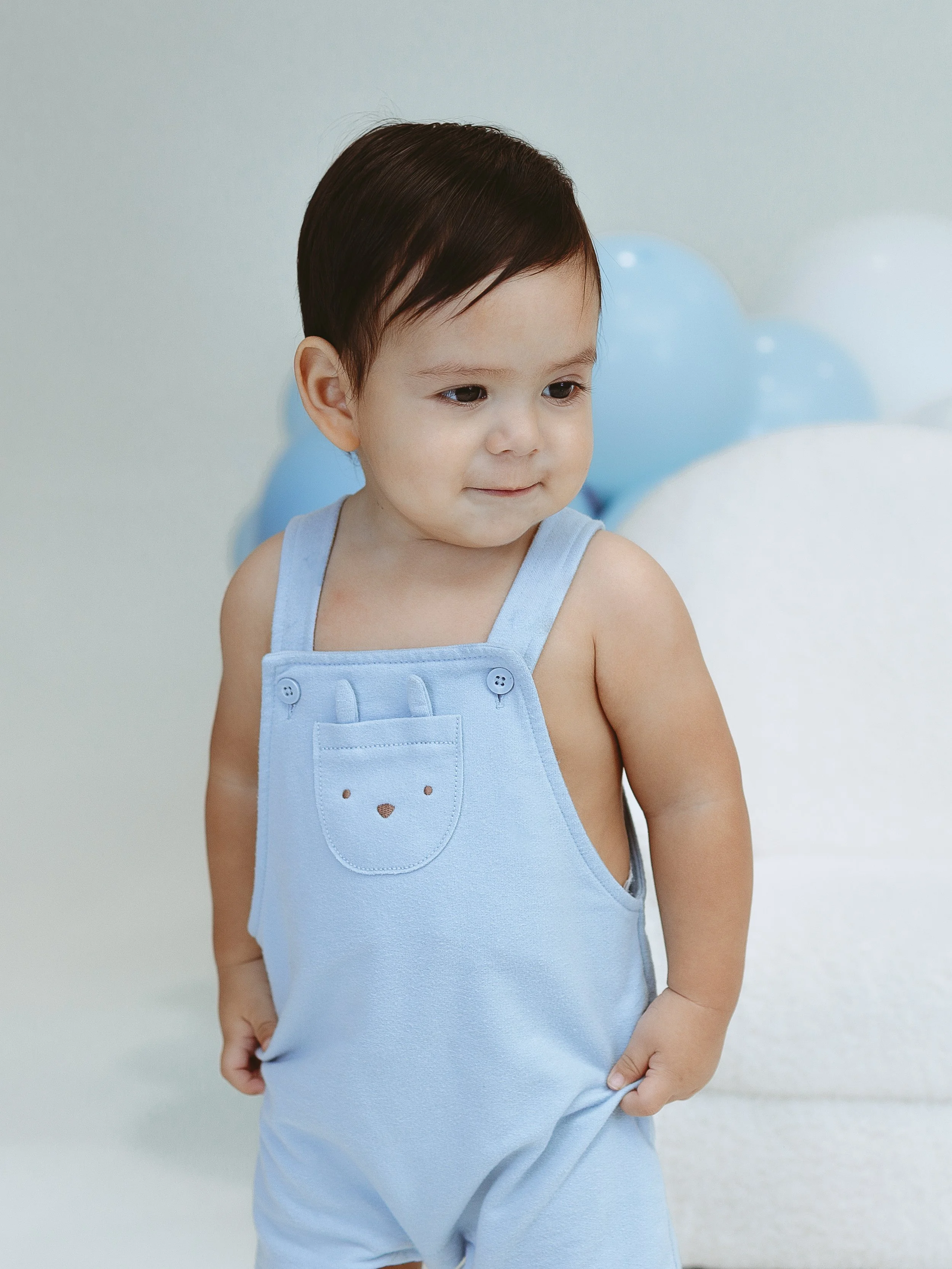 A young boy in a light blue sleeveless romper with a bear face pocket, standing in front of blue and white balloons.