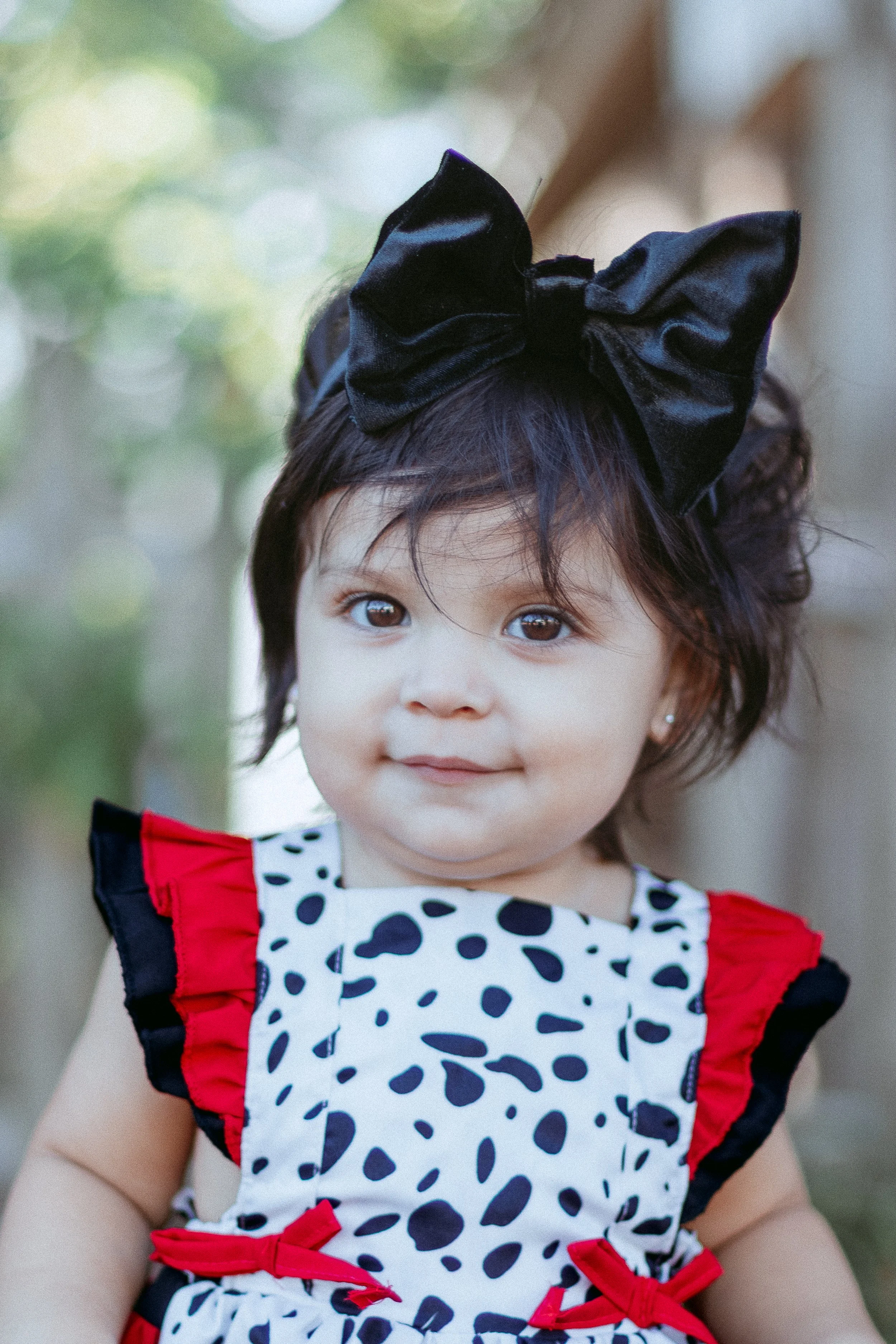 A young girl with dark hair, wearing a large black bow headband and a white dress with black and red accents and a black and white spotted pattern.