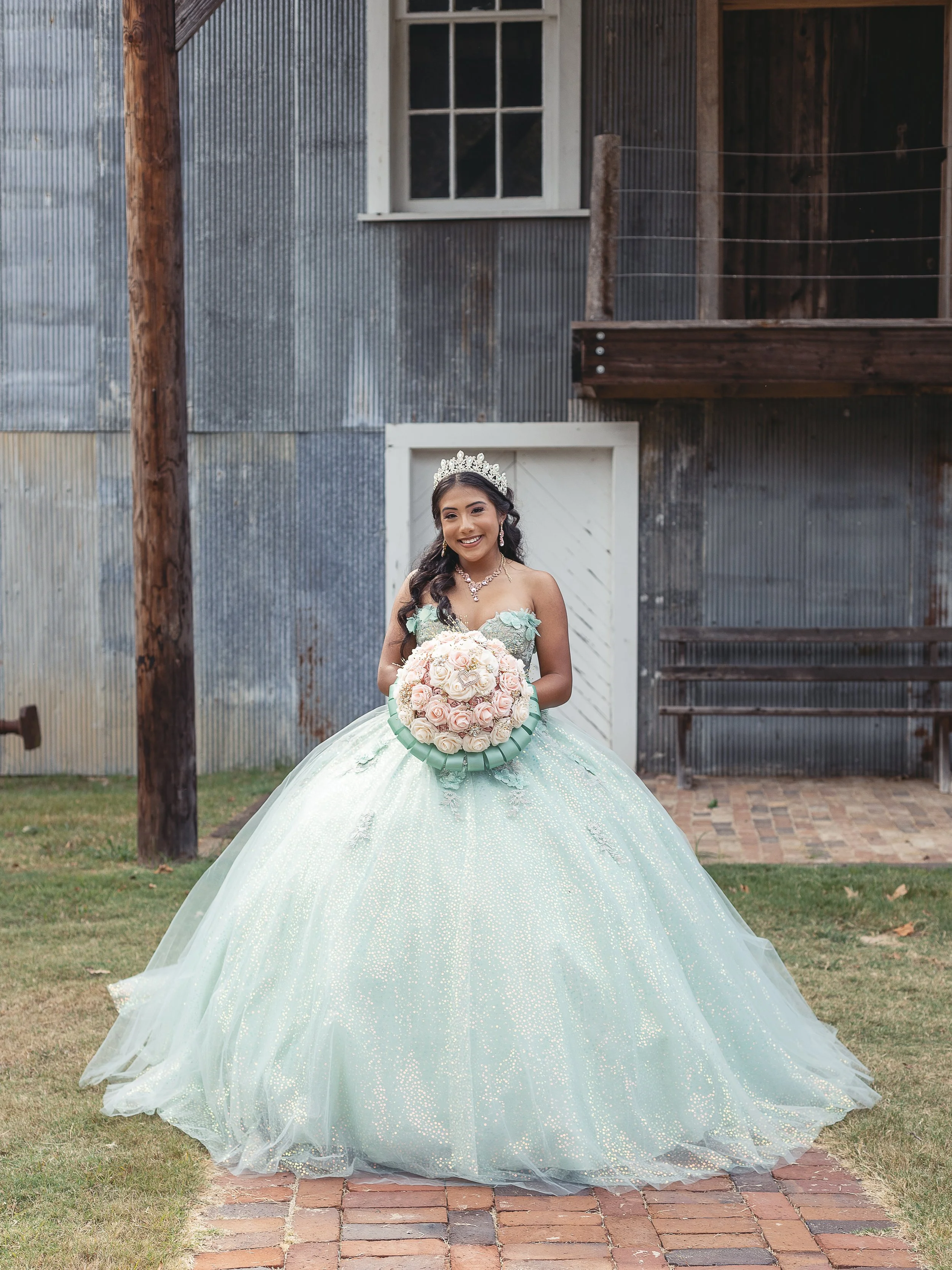 Young woman in a light green, sparkly ball gown with off-shoulder sleeves, wearing a tiara and necklace, stands outdoors holding a bouquet of pink and white roses, smiling.