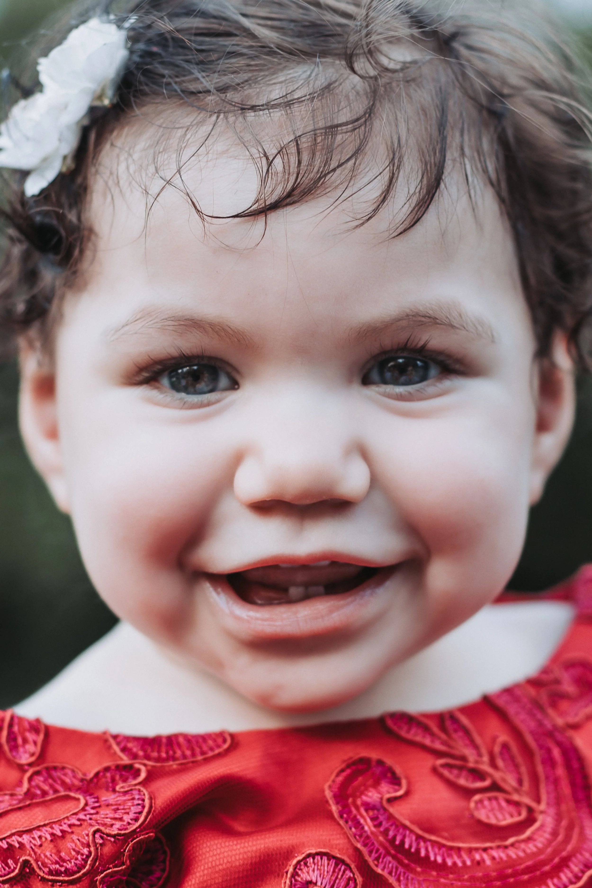 Close-up of a smiling young girl with curly brown hair, blue eyes, wearing a red dress with pink embroidery, and a white floral hair accessory.