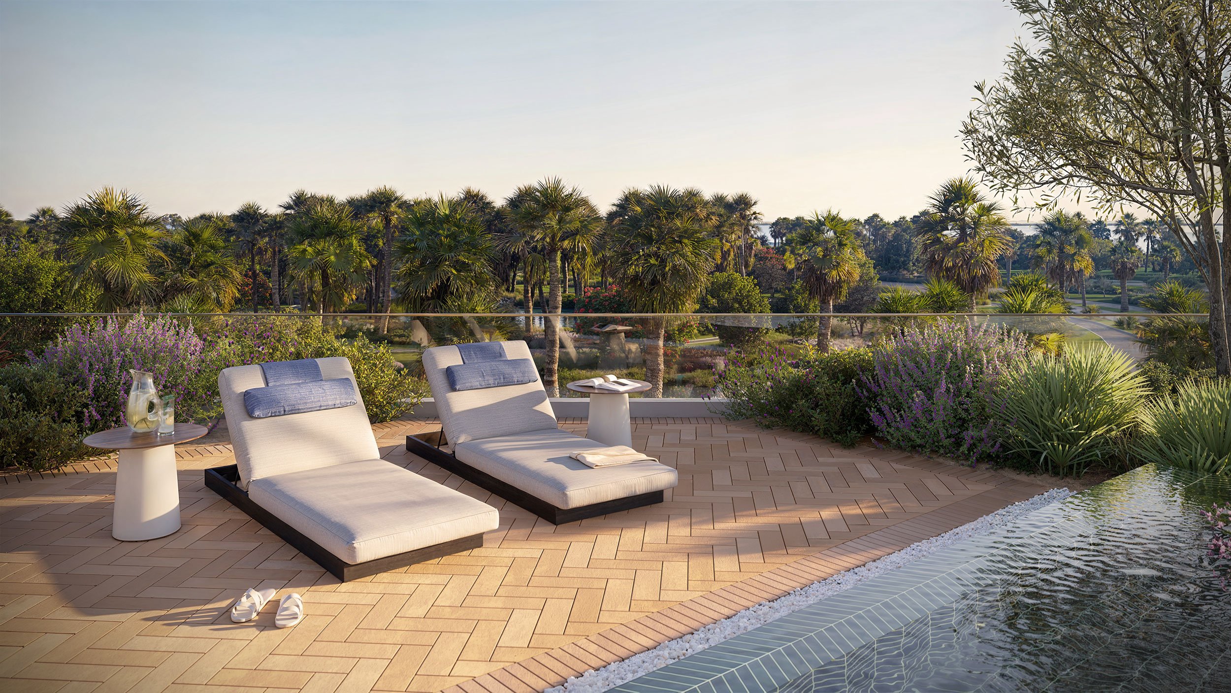 Luxury outdoor patio with two cream-colored lounge chairs, side tables, and a pool, overlooking a lush landscape of palm trees and flowering bushes.  Beyond Properties. Dubai Real Estate Marketing.