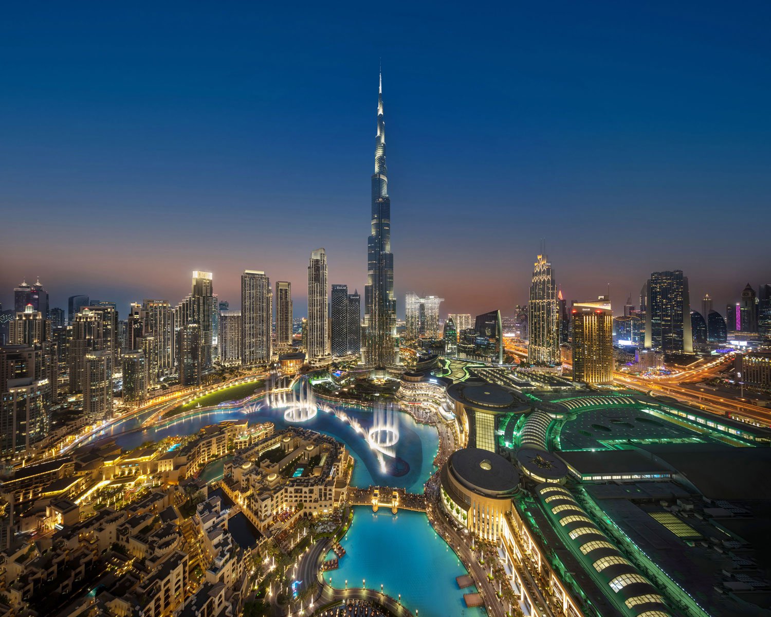 Night cityscape of Dubai featuring the Burj Khalifa, illuminated buildings, artificial lakes with fountains, and busy streets with lights.