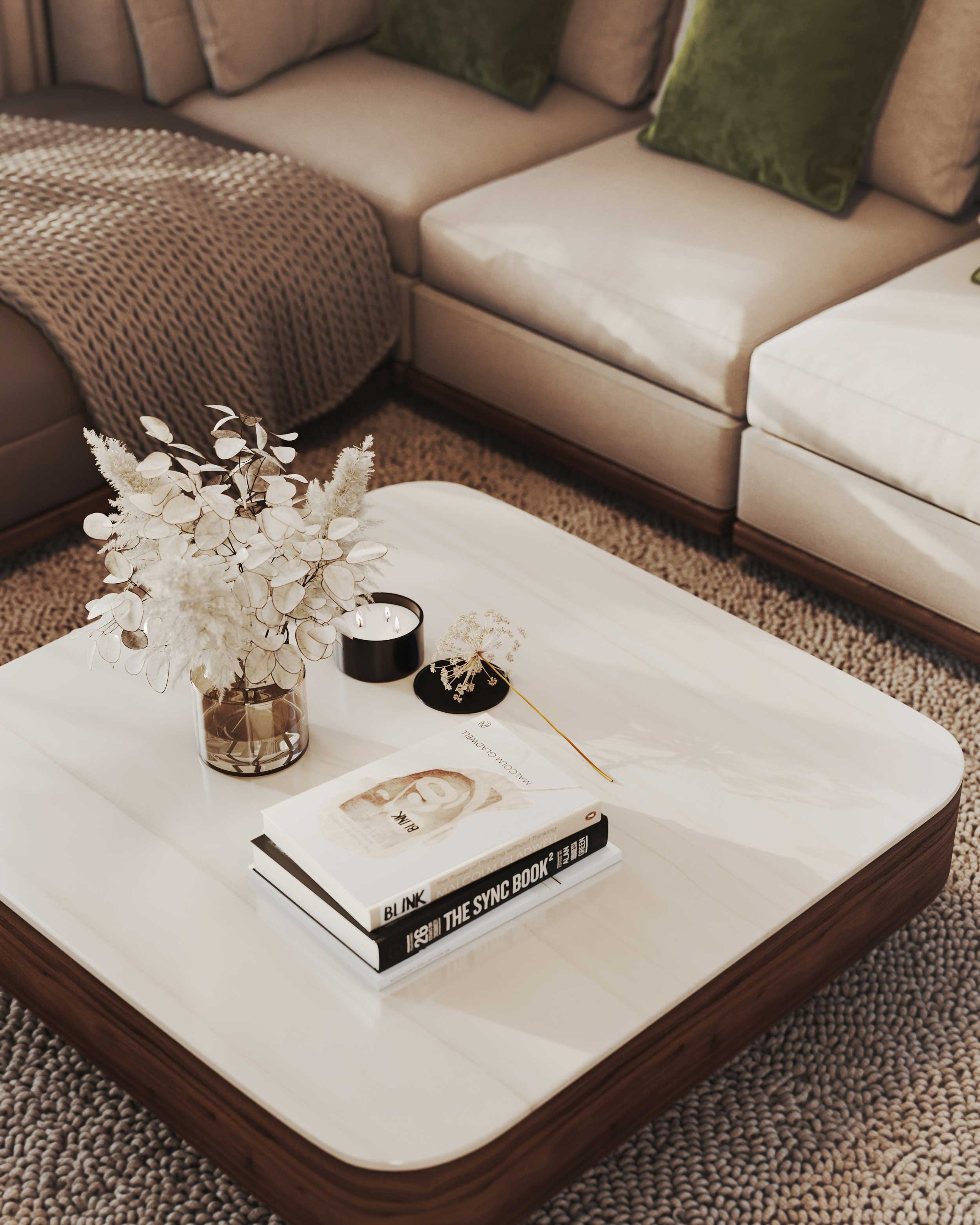 Living room with a white coffee table holding a book, a magazine, a small bouquet, and candles, with a gray L-shaped sofa and brown and green cushions.