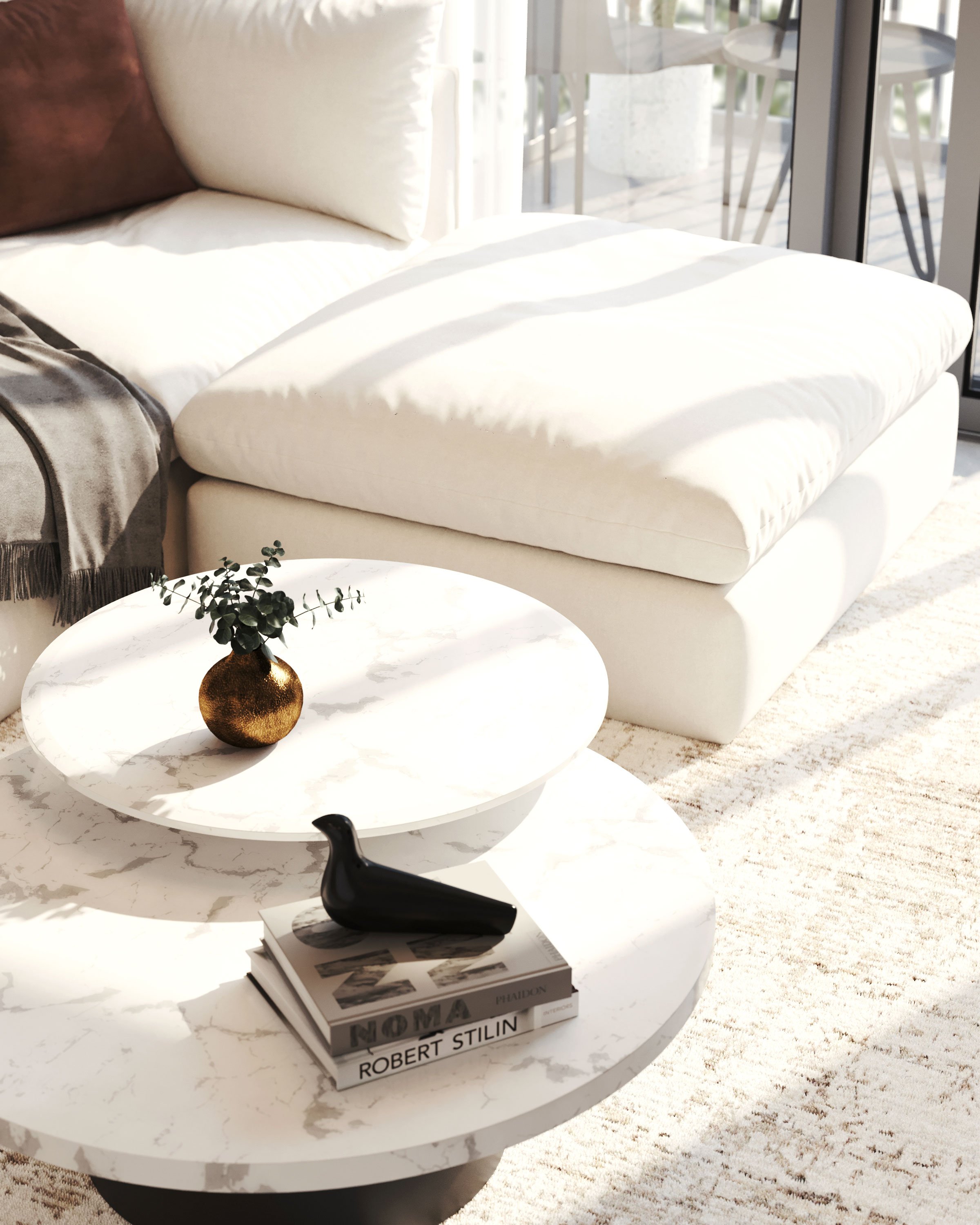 Living room with a white sectional sofa, a round marble coffee table with a small gold vase and green leaves, and a stack of books including 'NOMA' and 'Robert Stilin' with a black bird figurine on top. There is sunlight creating shadows on the sofa 