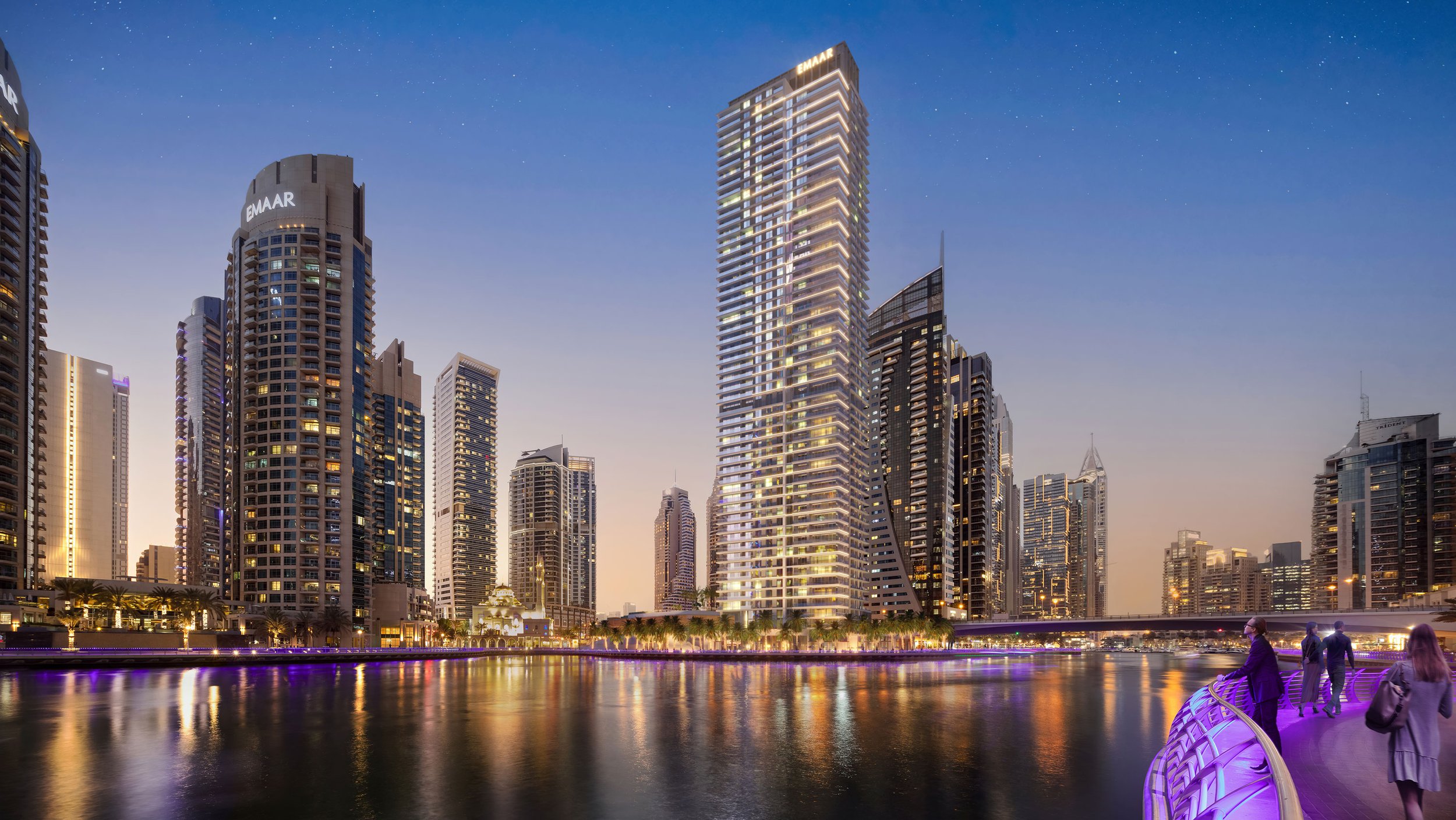 Night view of a city skyline with tall modern skyscrapers illuminated, reflected in a river, with a lit walkway and pedestrians in the foreground. Emaar. Dubai Marina. Dubai Real estate marketing.