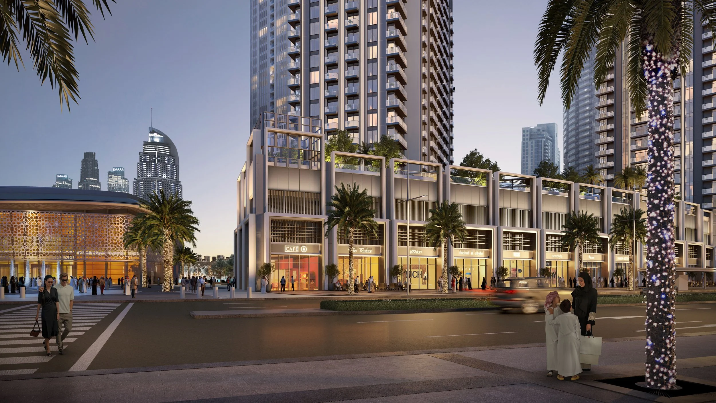 A modern cityscape at dusk featuring a multi-story commercial building with retail shops, surrounded by high-rise residential towers and skyscrapers in the background. Pedestrians cross the street, and palm trees line the sidewalk. Emaar. Dubai Downt