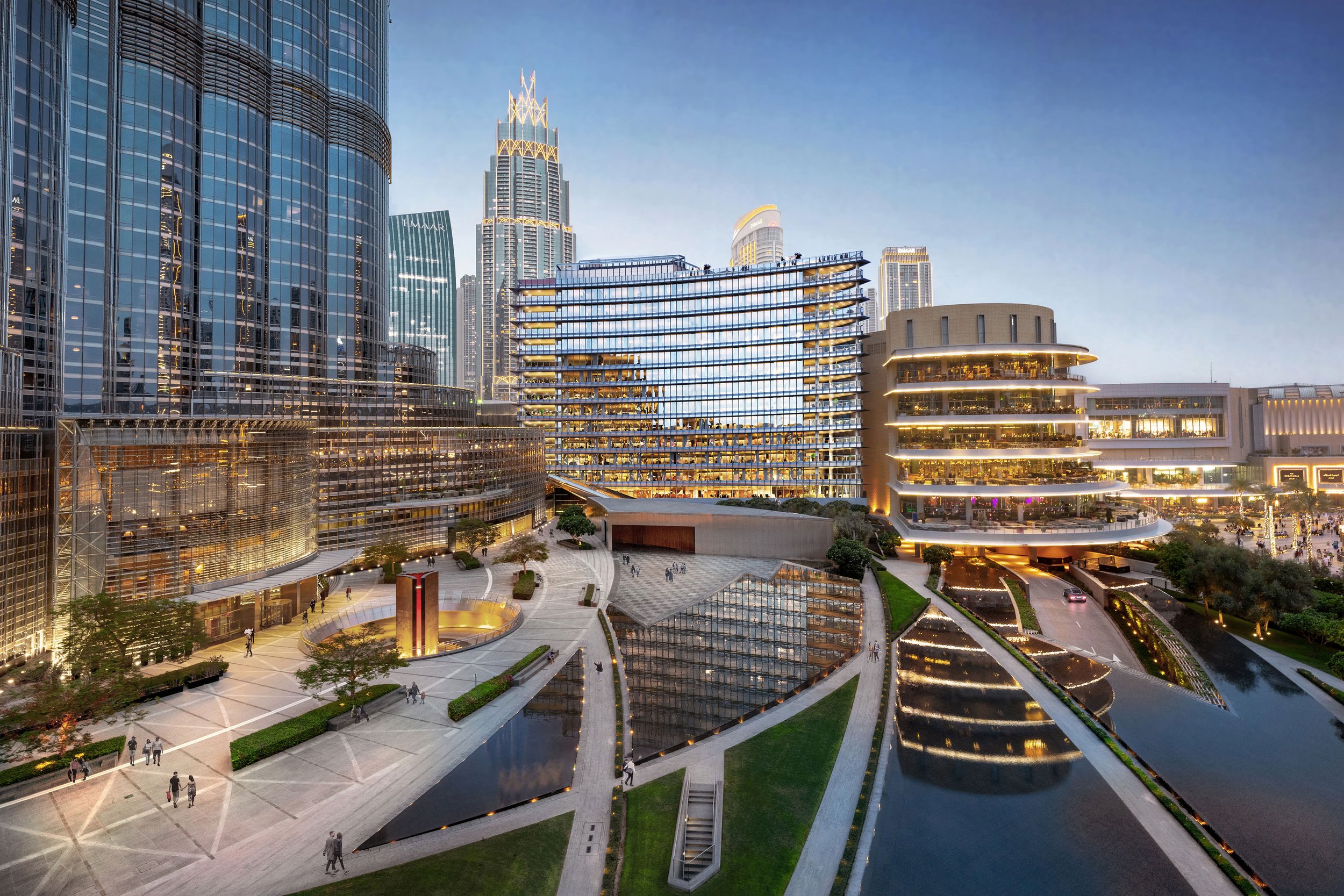 Modern cityscape with tall skyscrapers, a reflective glass building, plaza with trees and water features, and a lively commercial area, during dusk with illuminated buildings.