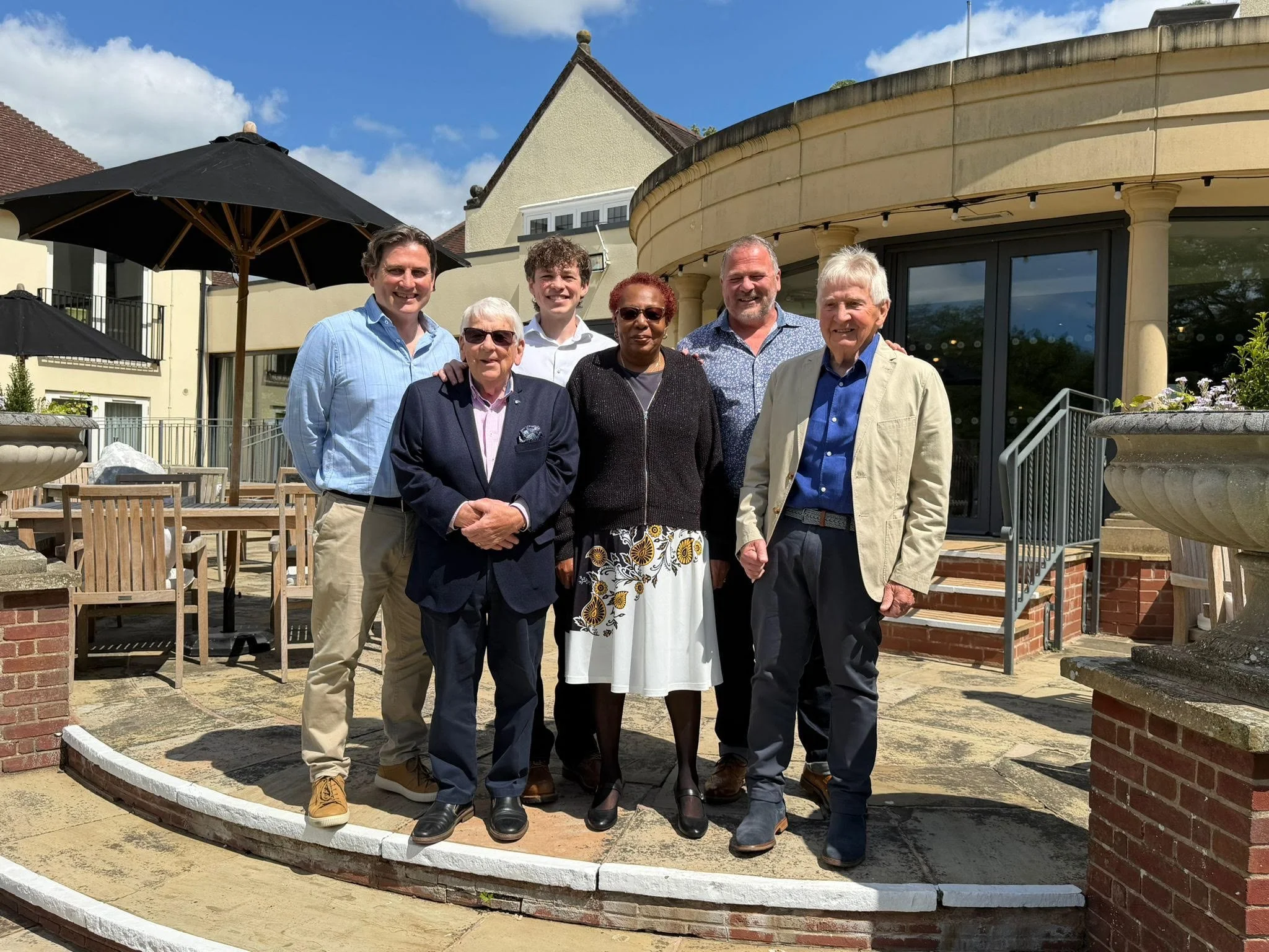 Group of seven people standing outdoors in front of a building, posing for a photo on a sunny day.
