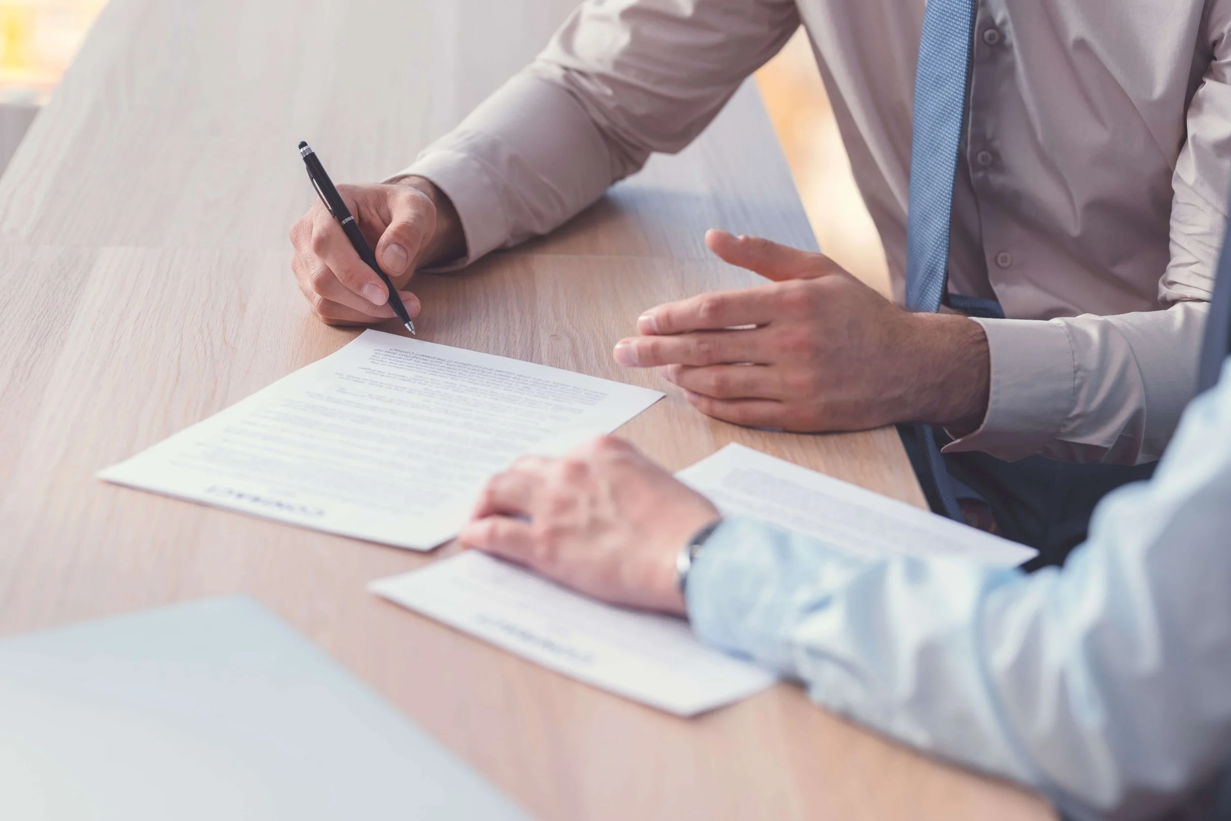 Close-up of two individuals in formal attire discussing documents on a desk, with one person holding a pen and the other gesturing with their hand.