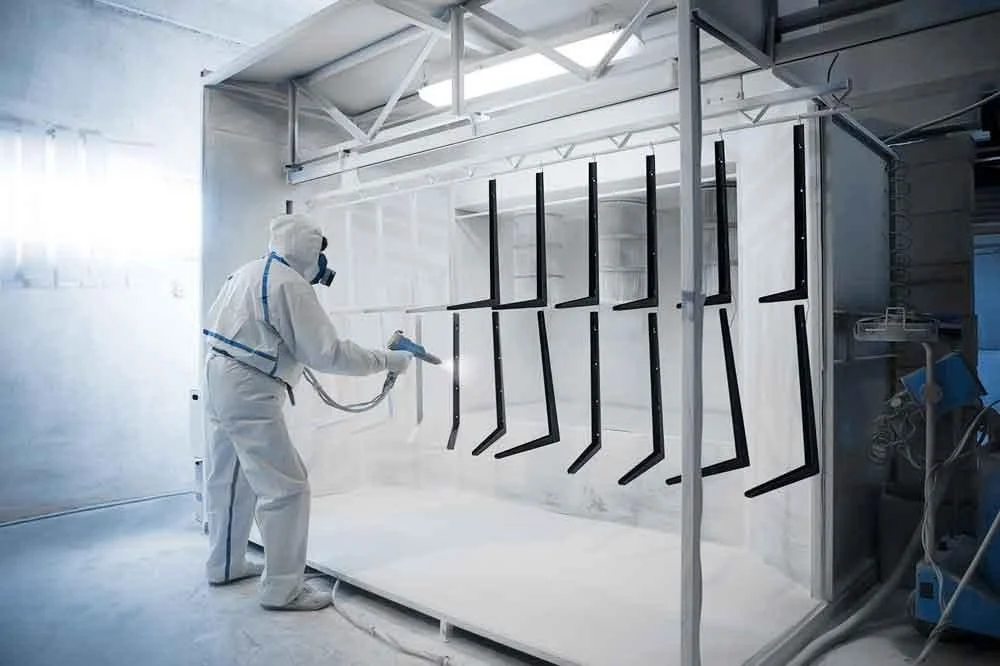 A person in protective gear spray paints metallic black legs of chairs inside a white, enclosed spray booth.