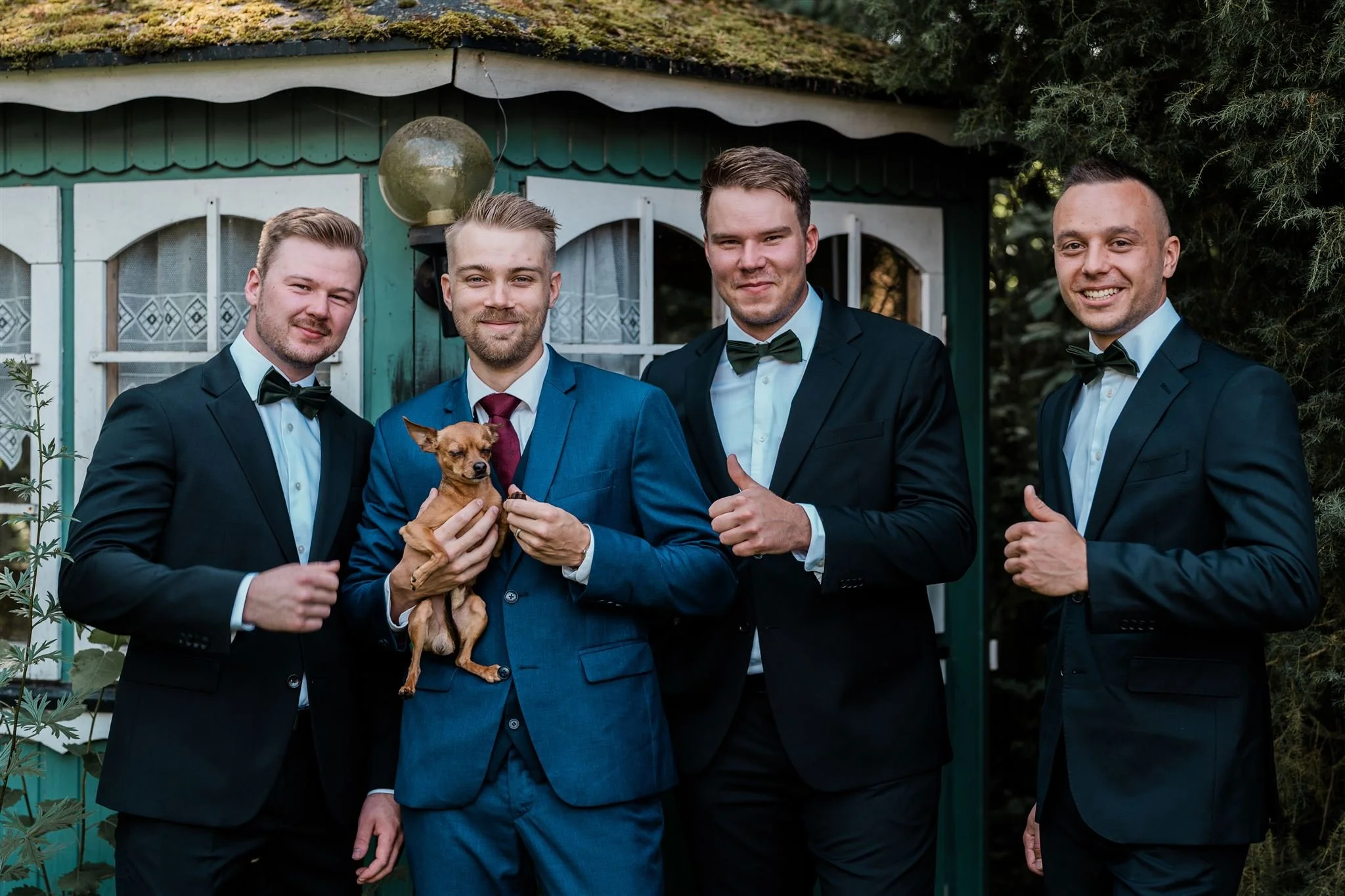 Neljän miehen ryhmä tunteellisissa pikkutakissa ja solmioissa tervehtii toisiaan ulkoalueella, ja yksi mies pitää pienen koiran sylissään.