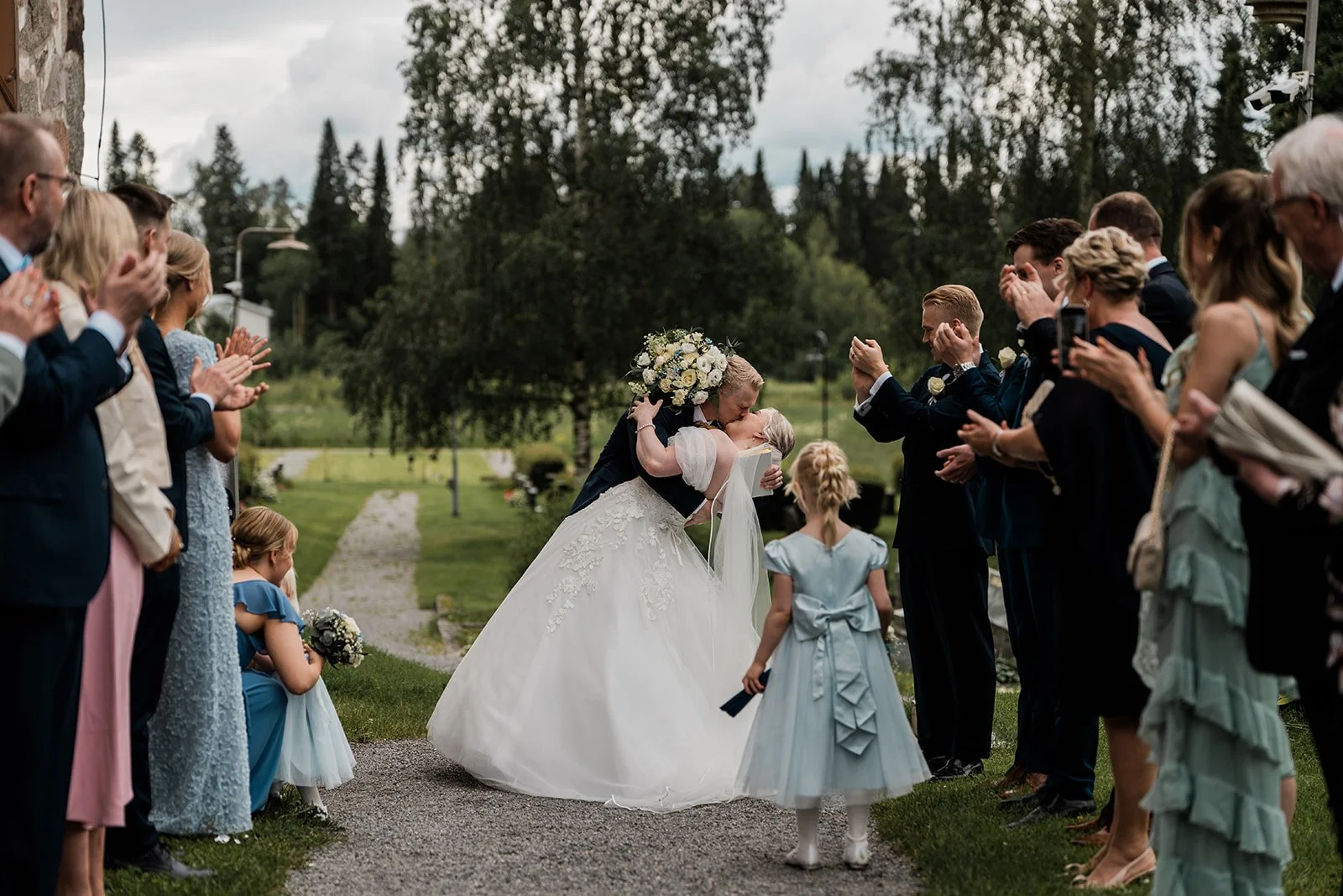 Hääpari suutelee toisinaan riviin asettuneiden vieraiden keskellä, vihkiseremonia ulkona, päiväsaikaan, vihkialueen ympärillä vihreää nurmikkoa ja puita, pilvinen taivas.