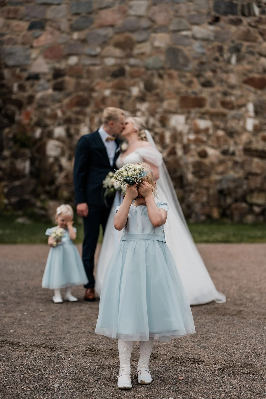 Lapsi peittää kasvonsa kukkakimpulla edessä, taustalla hääpari suutelee ja toinen lapsi.