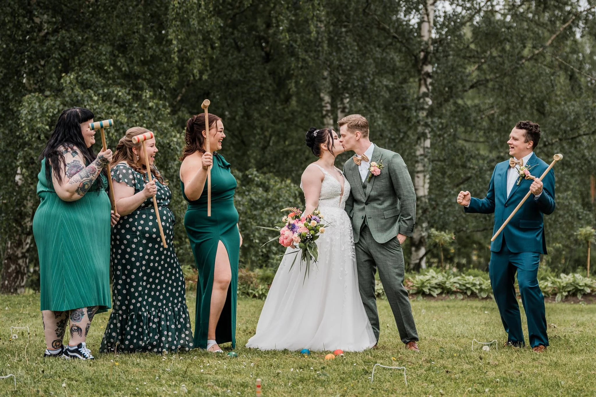 Hääpari suutelee toisinaan vihkimistilaisuudessa, ympärillä ystäviä, kaikki hymyilevät ja nauravat, ulkona puut kasvavat taustalla.