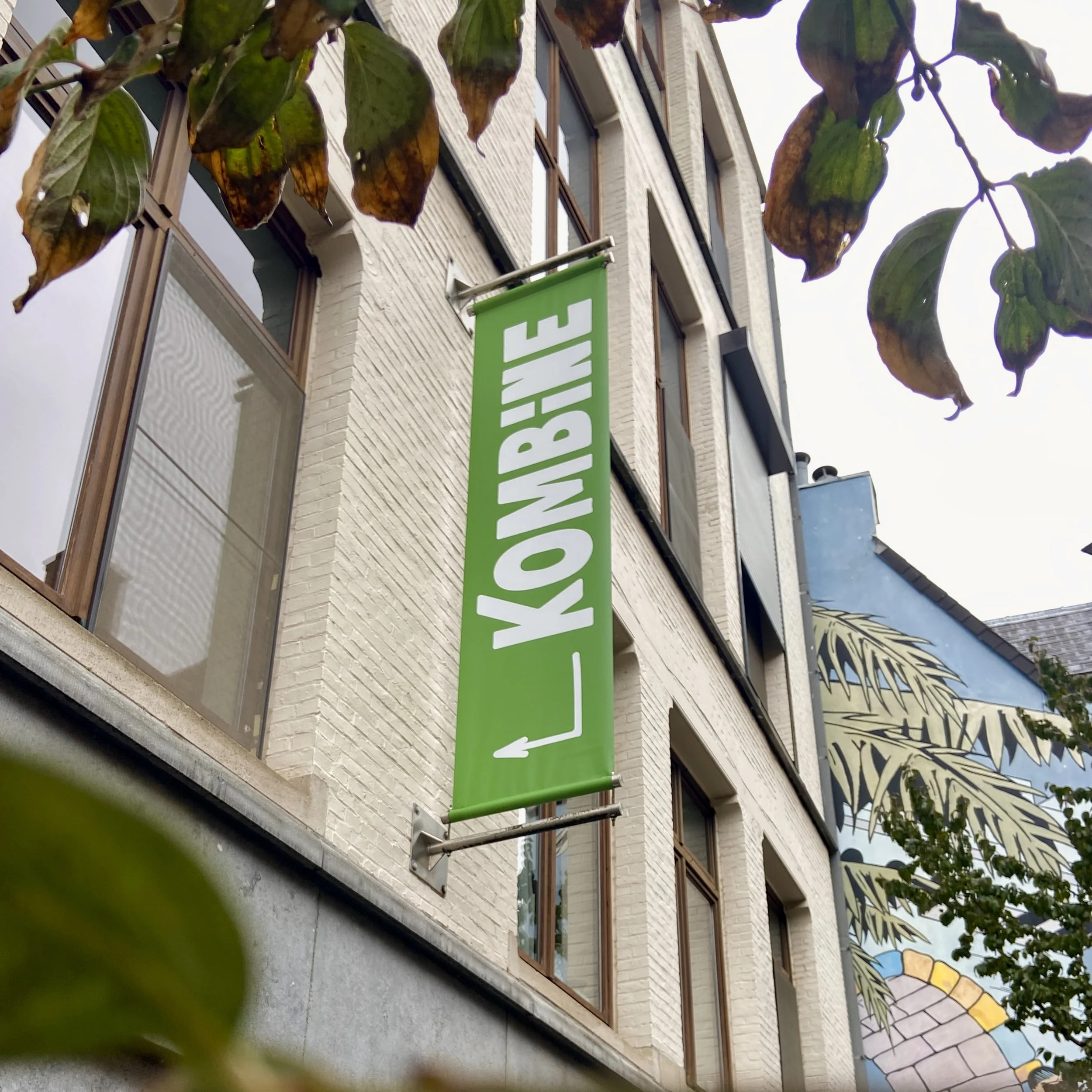 Groene verticale banner aan een gebouw met witte tekst die 'KOMBIJE' (ondersteboven) en een pijl aangeeft.