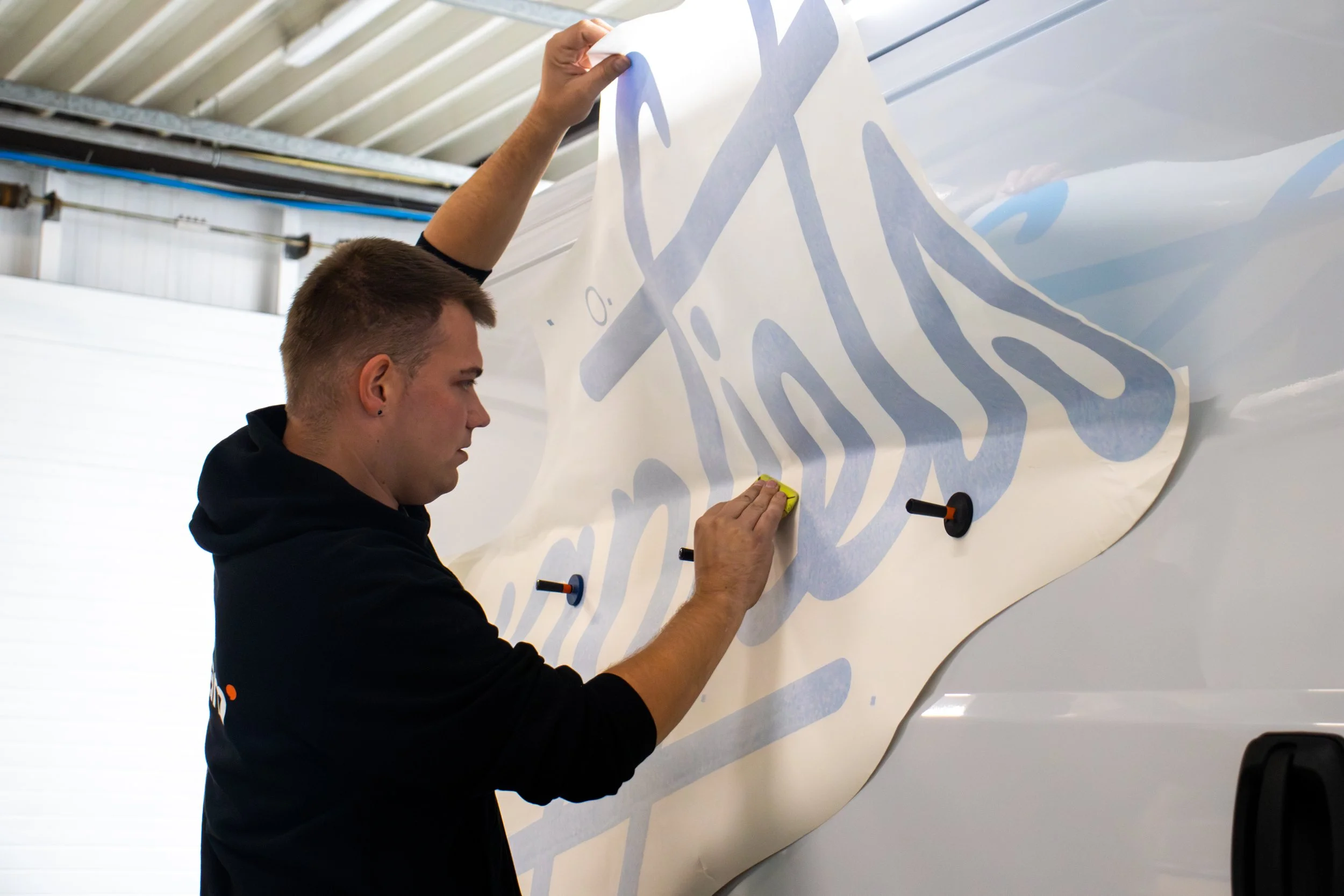Man plakt een grote sticker met blauwe tekst op de zijkant van een wit voertuig binnen een werkplaats.