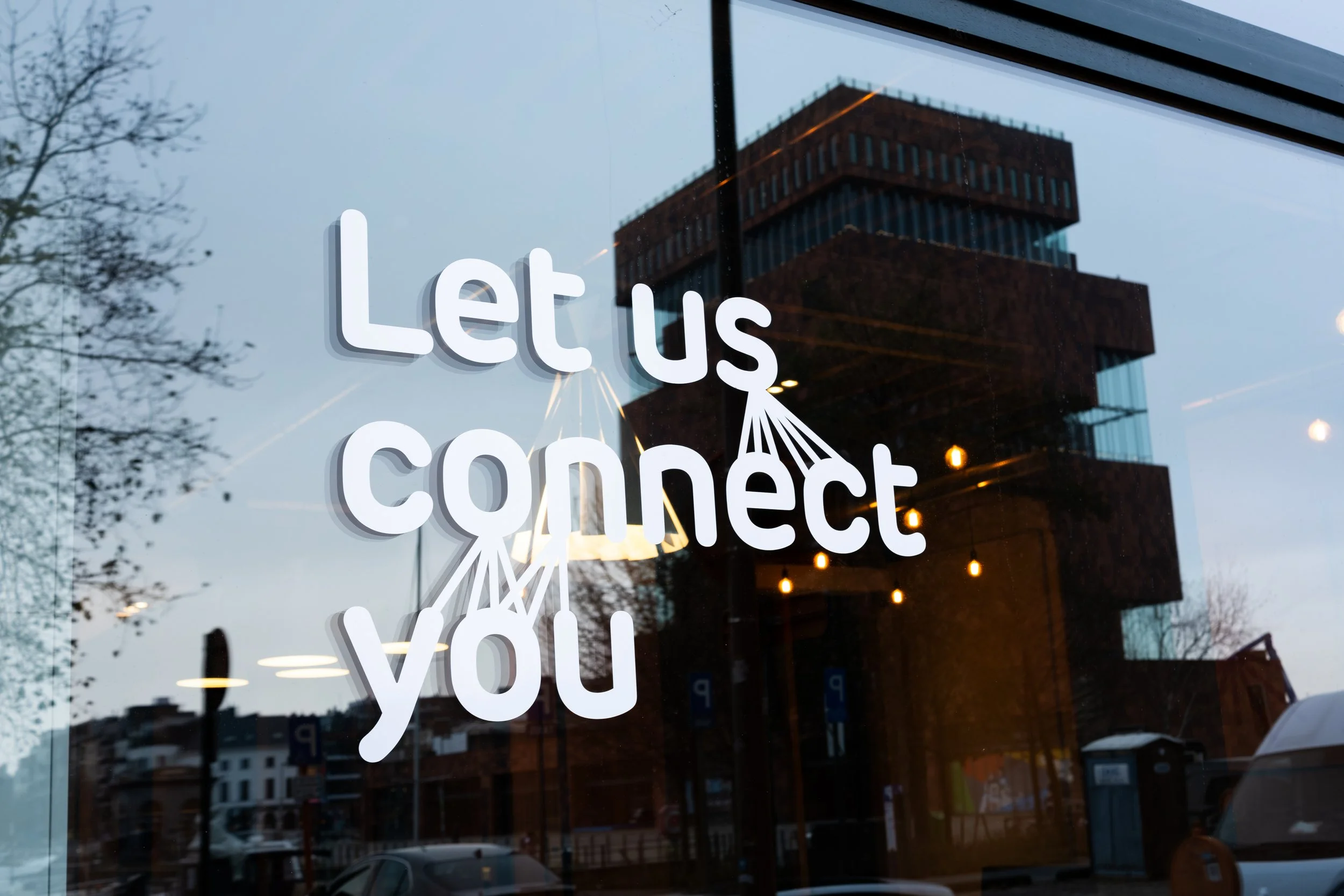 Tekst op een raam die zegt 'Let us connect you', met een reflectie van een gebouw en een lampen in de achtergrond.
