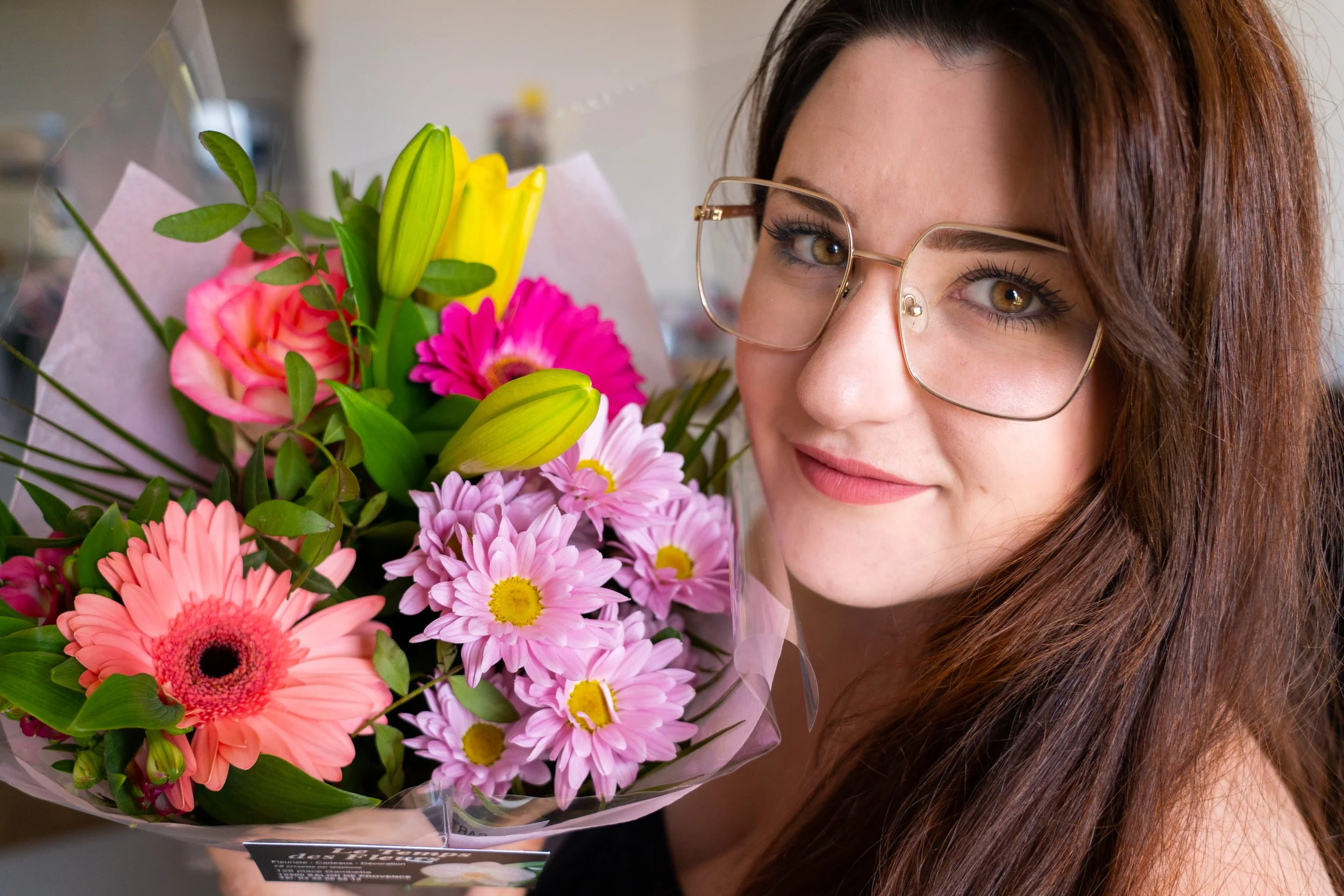 Femme souriante avec un bouquet de fleurs colorées, dont des gerberas et des marguerites, portant des lunettes à monture dorée.