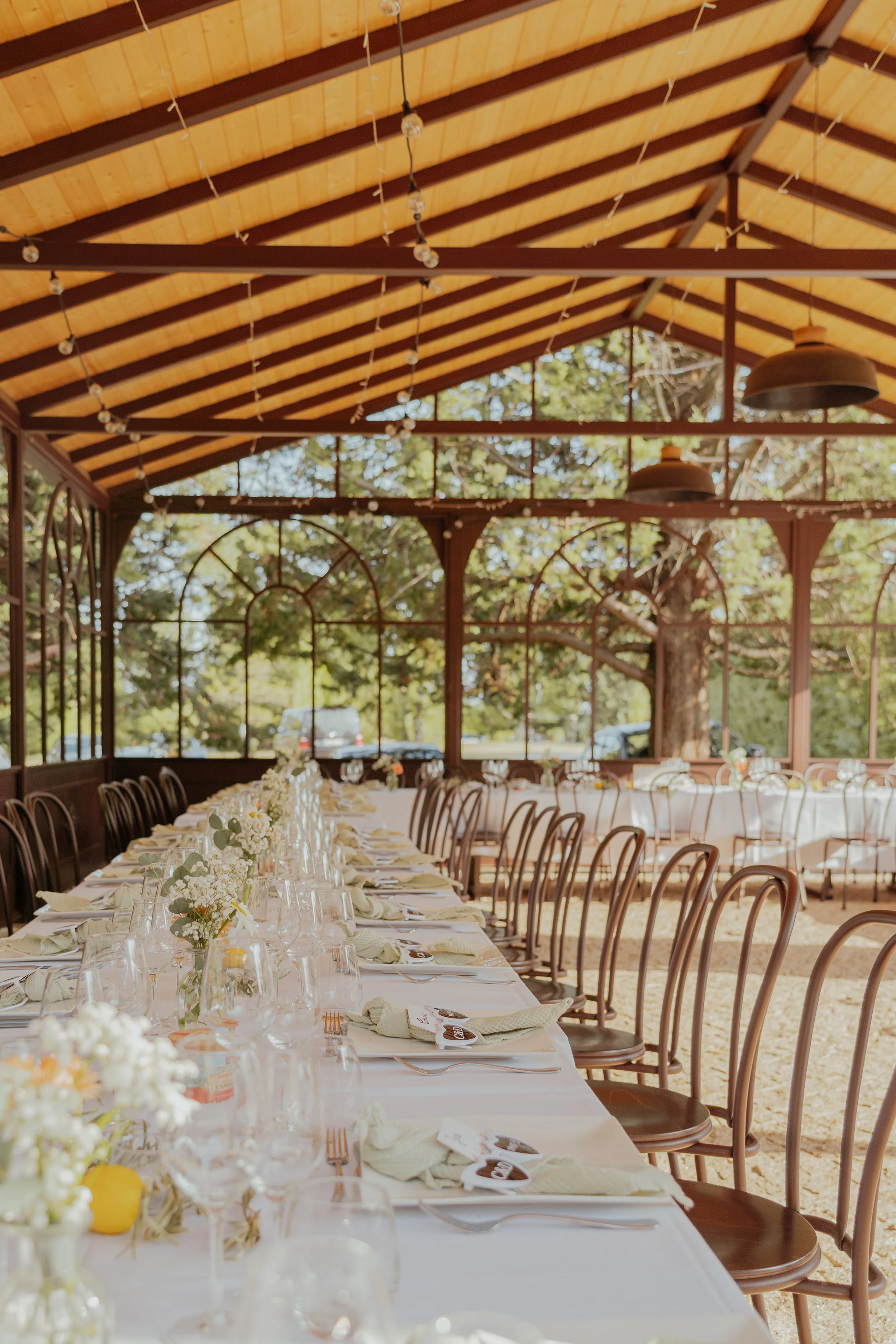 Une longue table de banquet décorée avec des arrangements floraux blancs, des verres à vin et des serviettes pliées, dans une tente en bois avec des murs en verre offrant une vue sur des arbres et des voitures à l'extérieur.