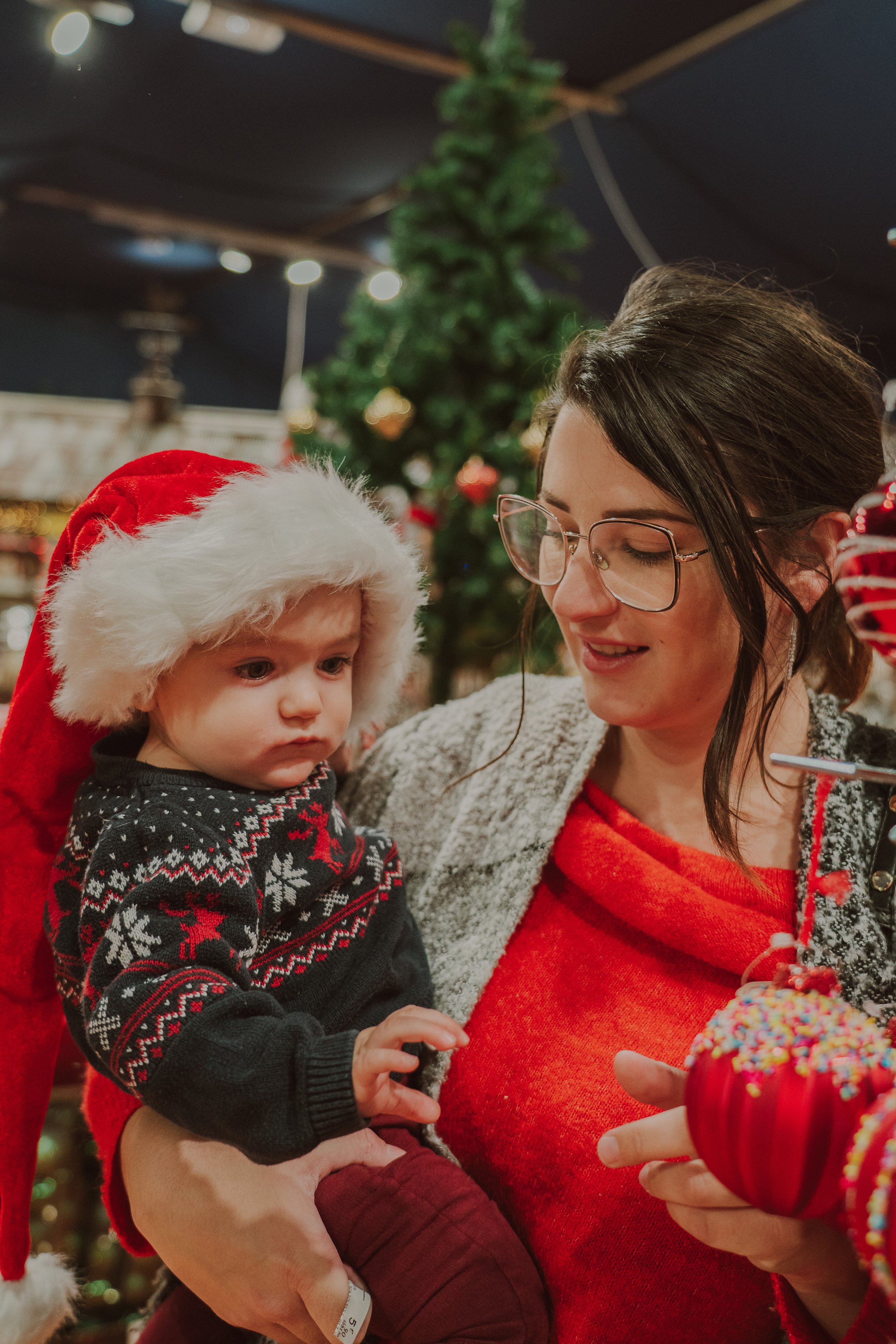 Une femme tient un bébé habillé en tenue de Noël, portant un bonnet de Père Noël, dans un décor festif avec un sapin de Noël en arrière-plan.