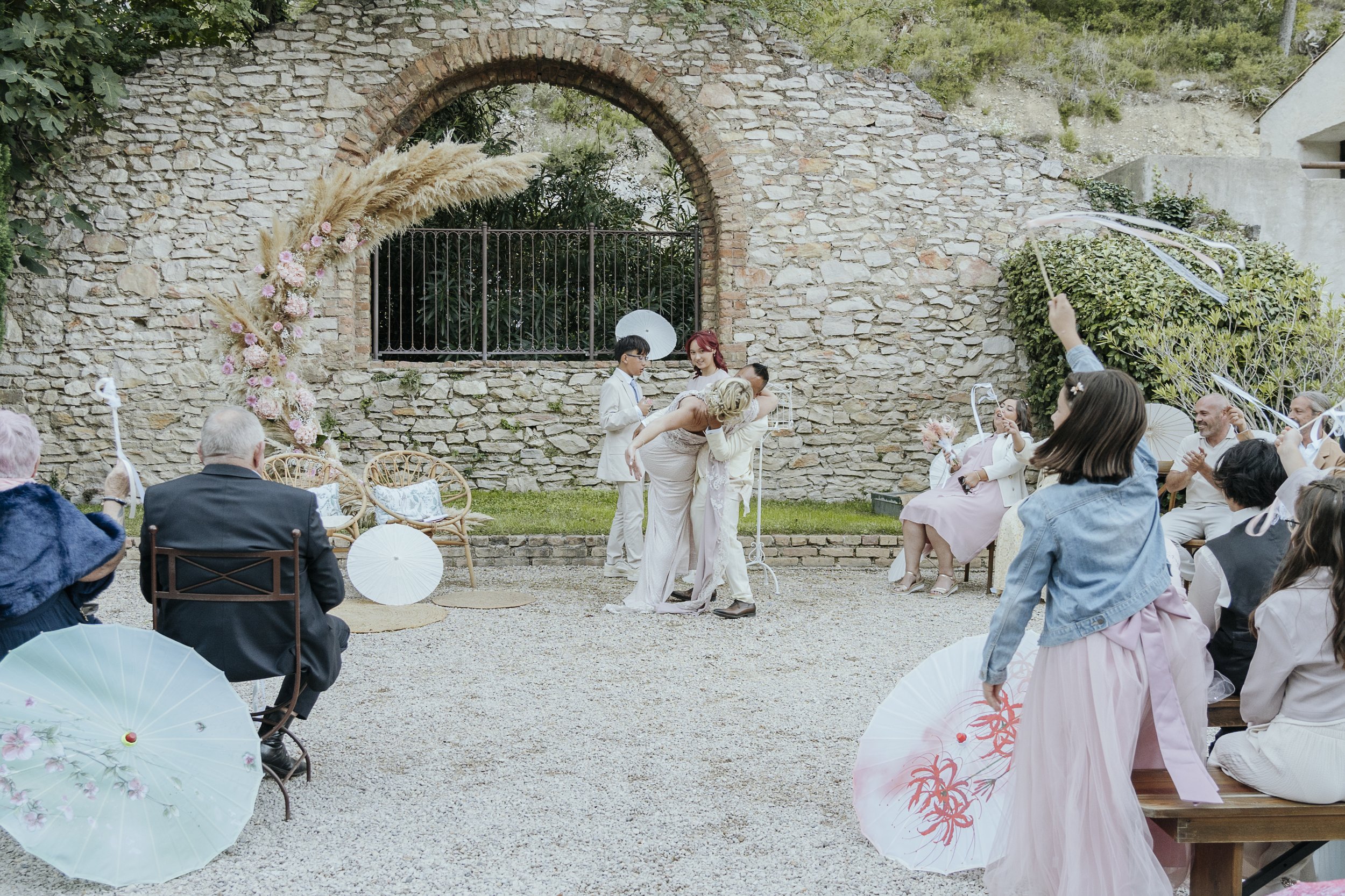 Une cérémonie de mariage en plein air devant un mur en pierre avec un arc décoré de fleurs et de pampas, invités assis sous des parapluies colorés, mariés échangeant un baiser, quelques personnes avec des parasols, ambiance joyeuse et festive.