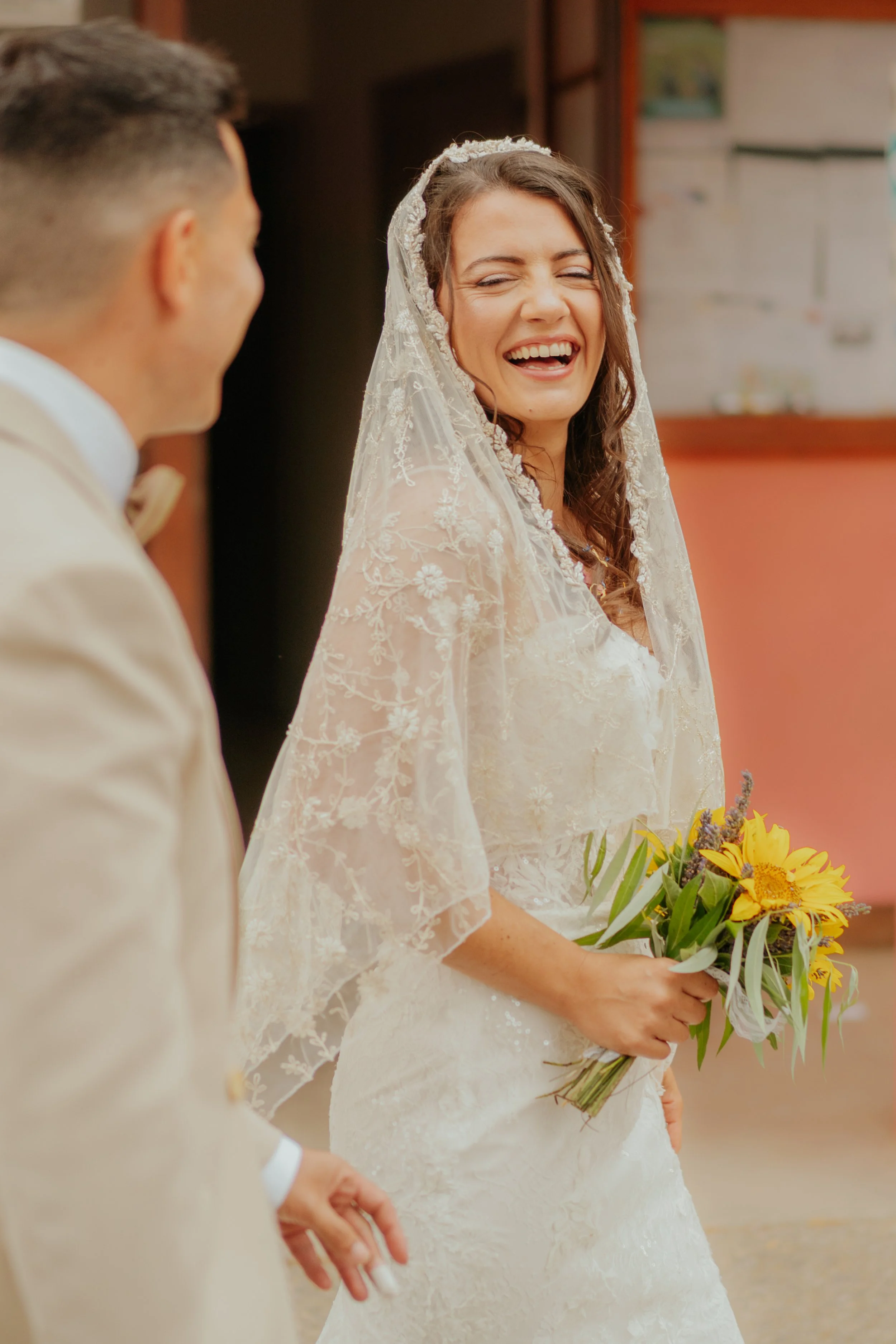 Une femme souriante en robe de mariage tenant un bouquet de tournesols, avec un homme en costume beige à côté.
