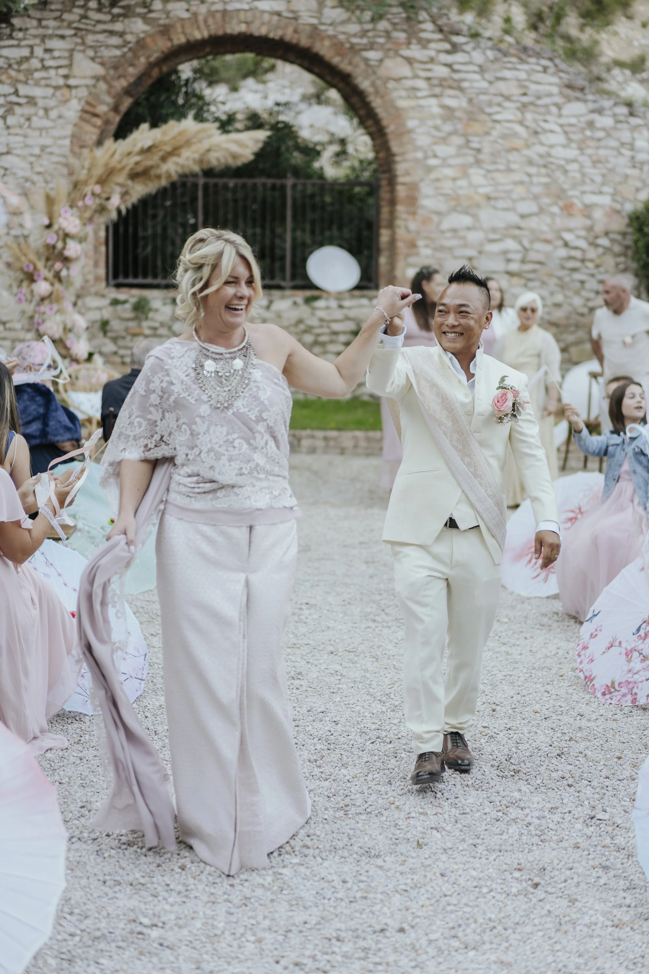 Un couple souriant, lors d'une cérémonie de mariage en plein air, entouré d'invités, avec une structure en pierre en arrière-plan.