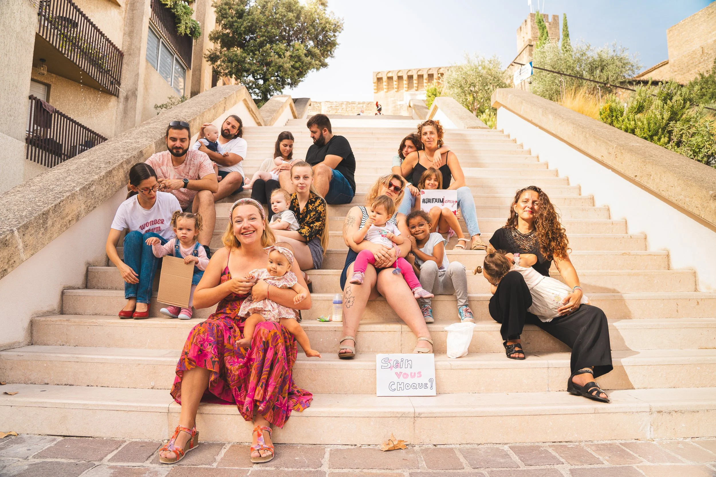 Un groupe de personnes assises sur un escalier, comprenant des adultes et des enfants, avec des pancartes portant des messages. En arrière-plan, un bâtiment et des arbres.