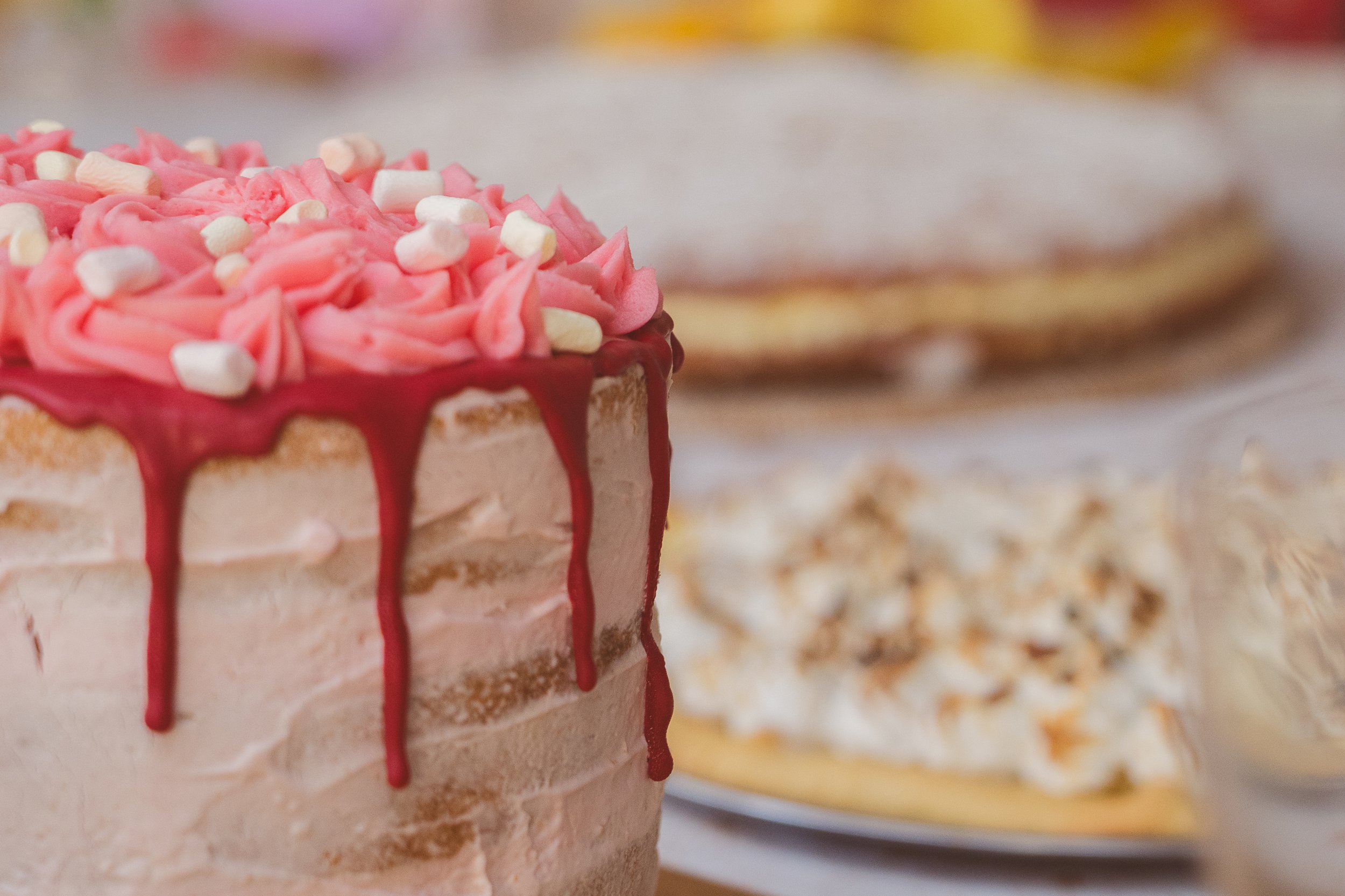 Gâteau décoré de glaçage rose avec des guimauves, nappé de coulis rouge, et d'autres desserts flous en arrière-plan.