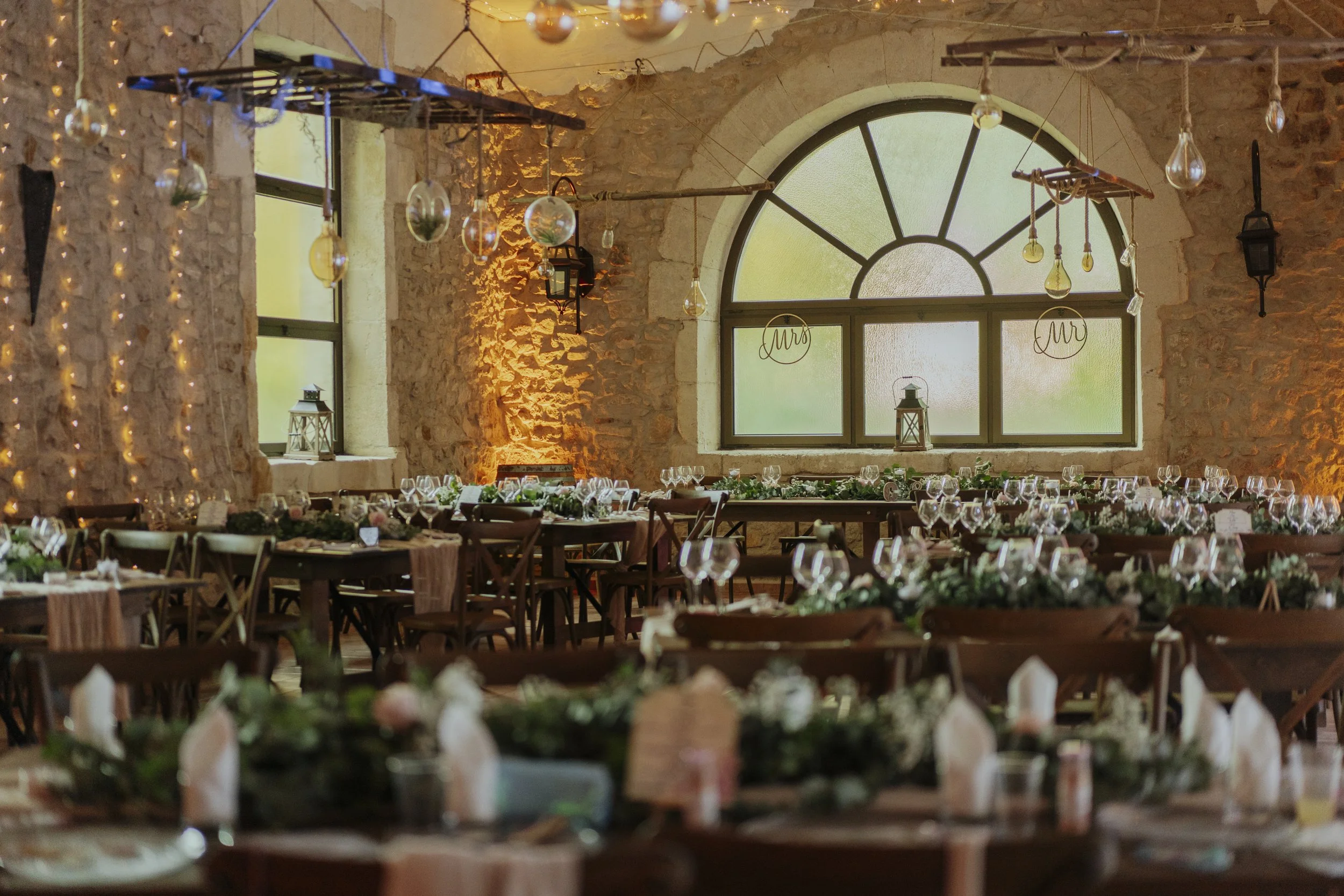 Salle de réception décorée pour un événement, avec grandes fenêtres, murs en pierre, suspensions de lumière, guirlandes lumineuses, et tables élégamment dressées avec vaisselle et fleurs.
