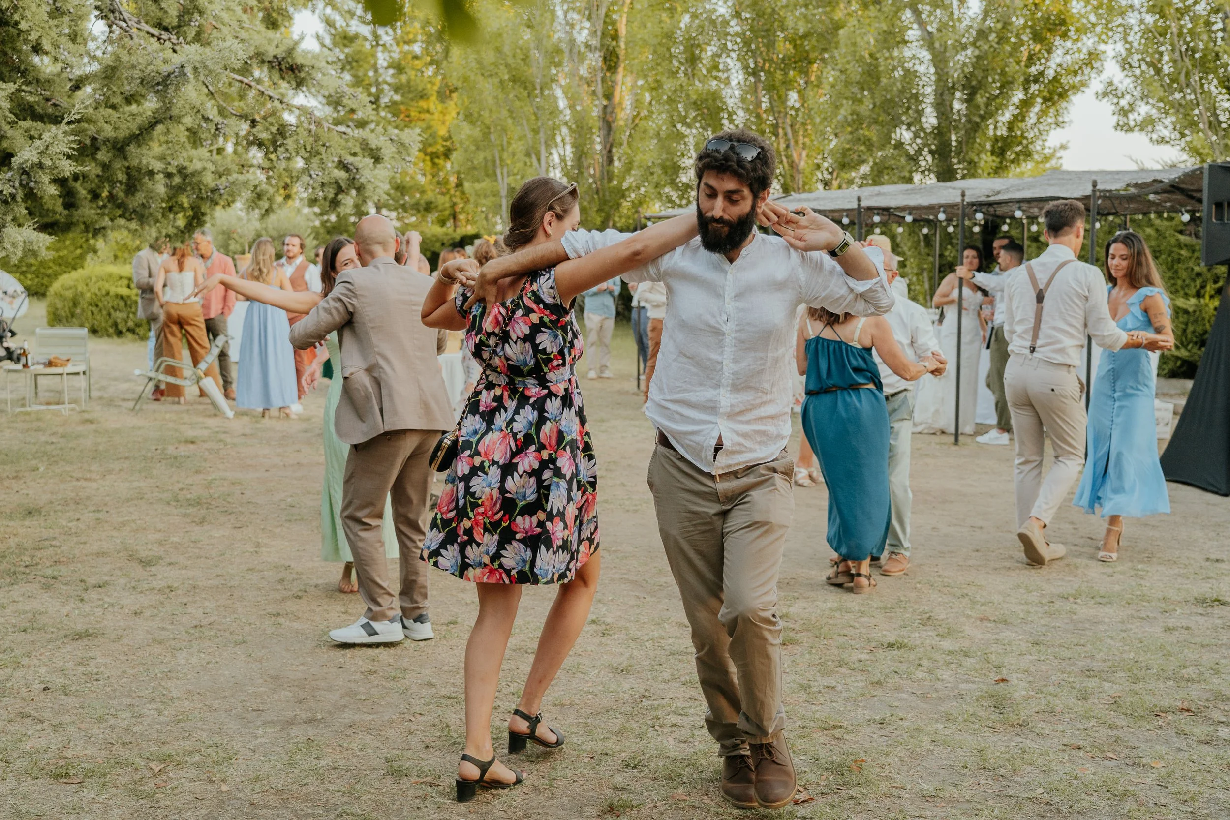 des personnes dansent lors d'une fête en plein air entourée d'arbres, portant des vêtements décontractés et colorés.