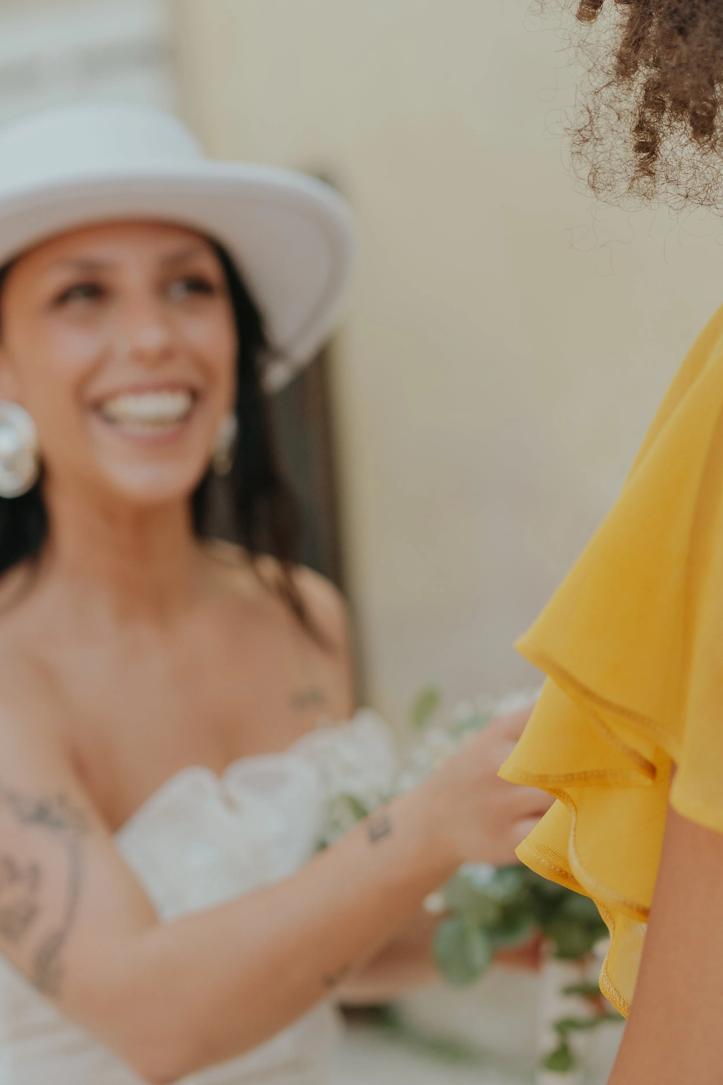 Une femme souriante portant un chapeau blanc et une robe blanche avec des fleurs, fête un mariage ou un événement spécial, face à une autre personne portant une robe jaune.