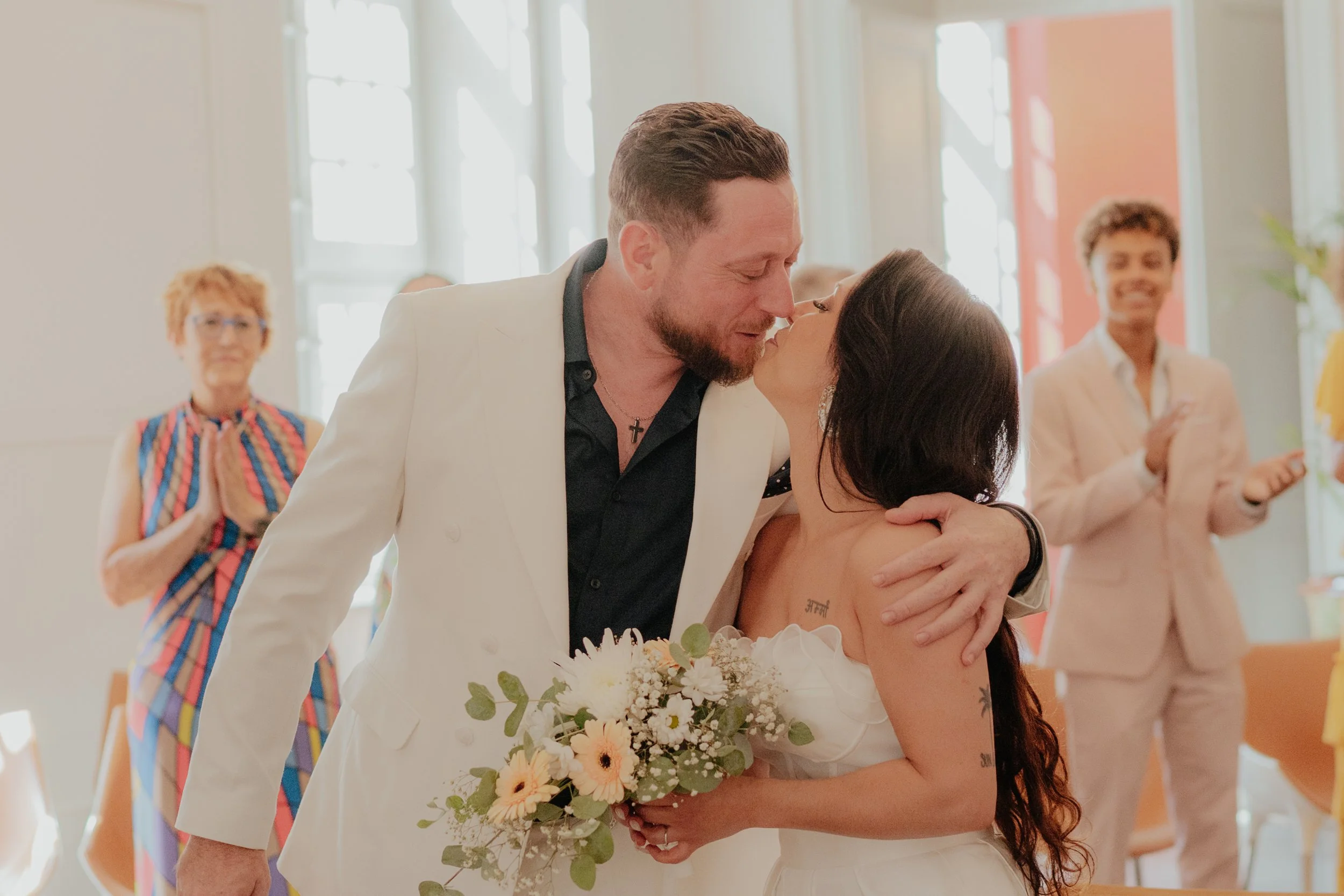 Un couple se embrasse lors d'un mariage, la femme tient un bouquet de fleurs et il y a des témoins en arrière-plan qui applaudissent, dans une salle lumineuse.