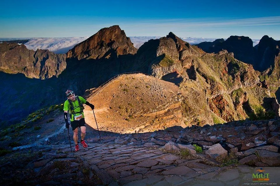 A MIUT runner with poles climbing a stone path through Madeira's central peaks, above a sea of cloud.