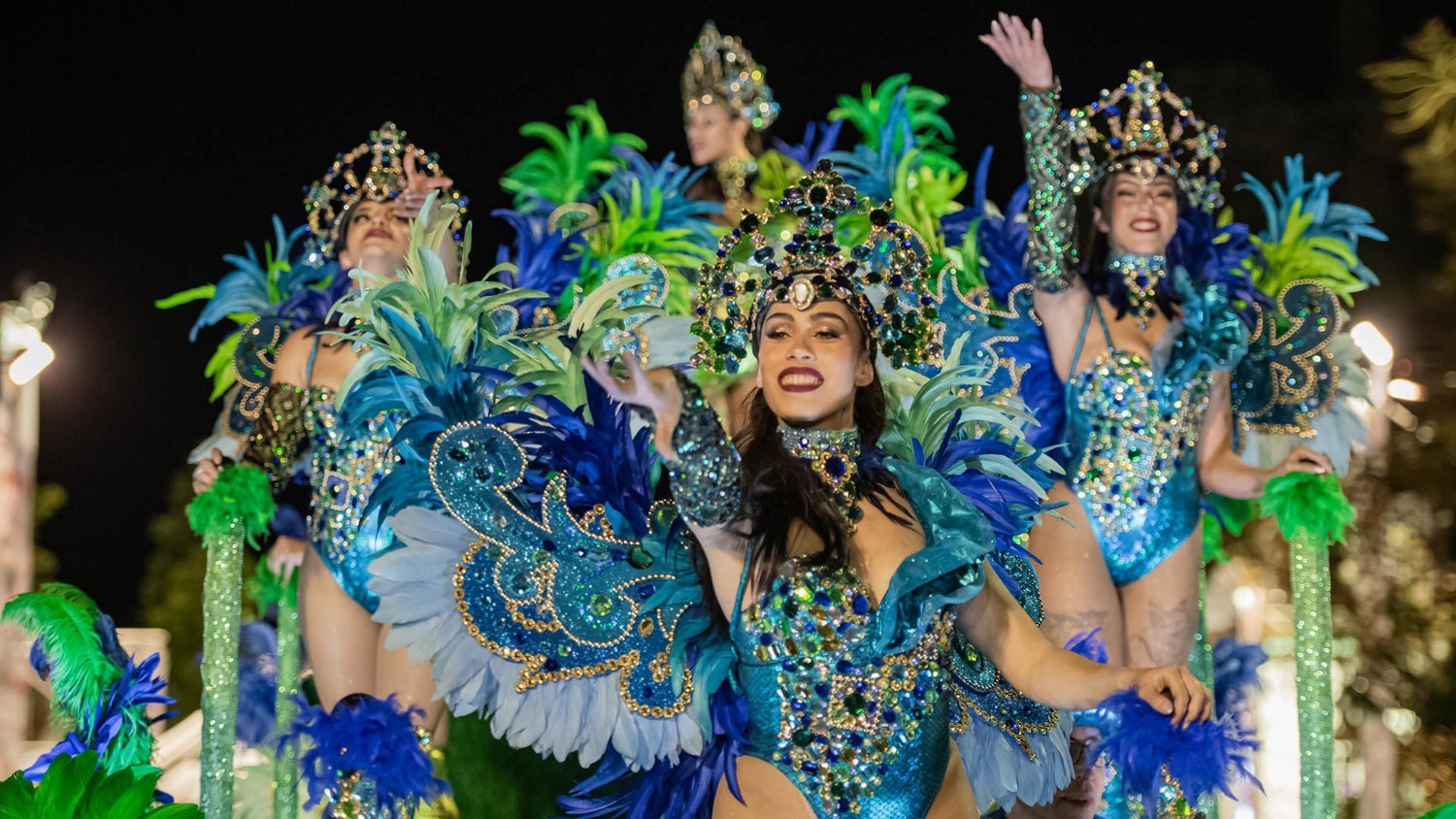 Samba dancers in blue and green feathered costumes on a lit float in the Madeira Carnival allegoric parade at night.