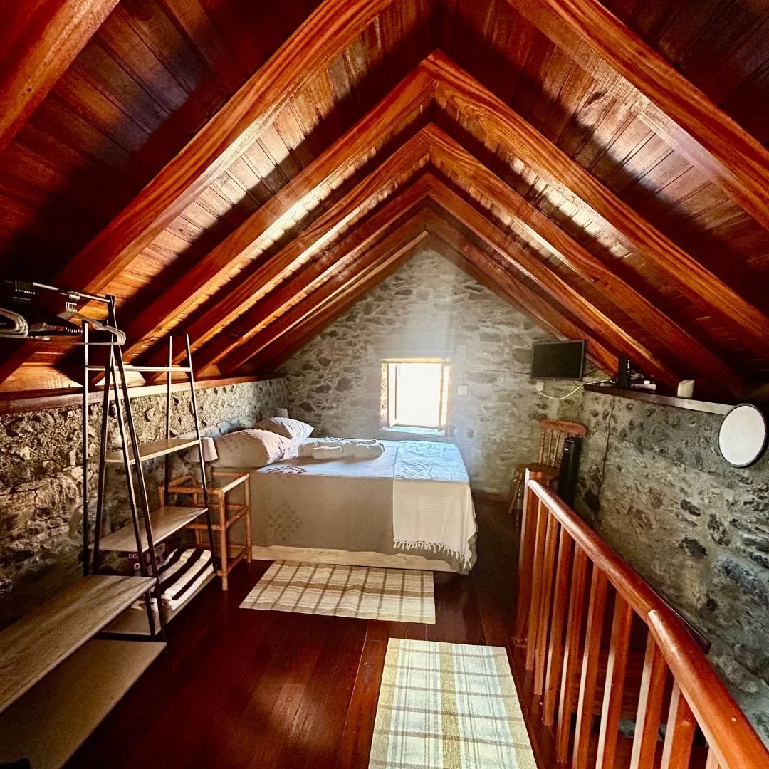 The sleeping loft under a full timber A frame of varnished beams, with a double bed against the stone gable and a small window above it