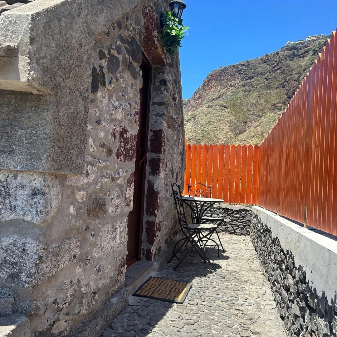 The cobbled path along the cottage with an iron table and chairs against the stone, an orange fence beside it and the green cliff rising behind