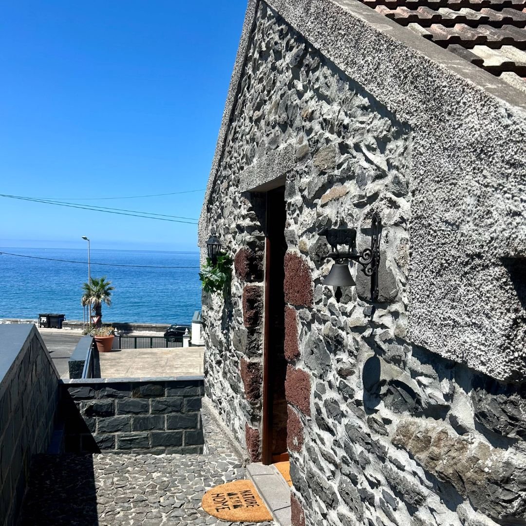 The cottage's open front door in a rough stone wall, with an iron bell beside it and the Atlantic across the road beyond