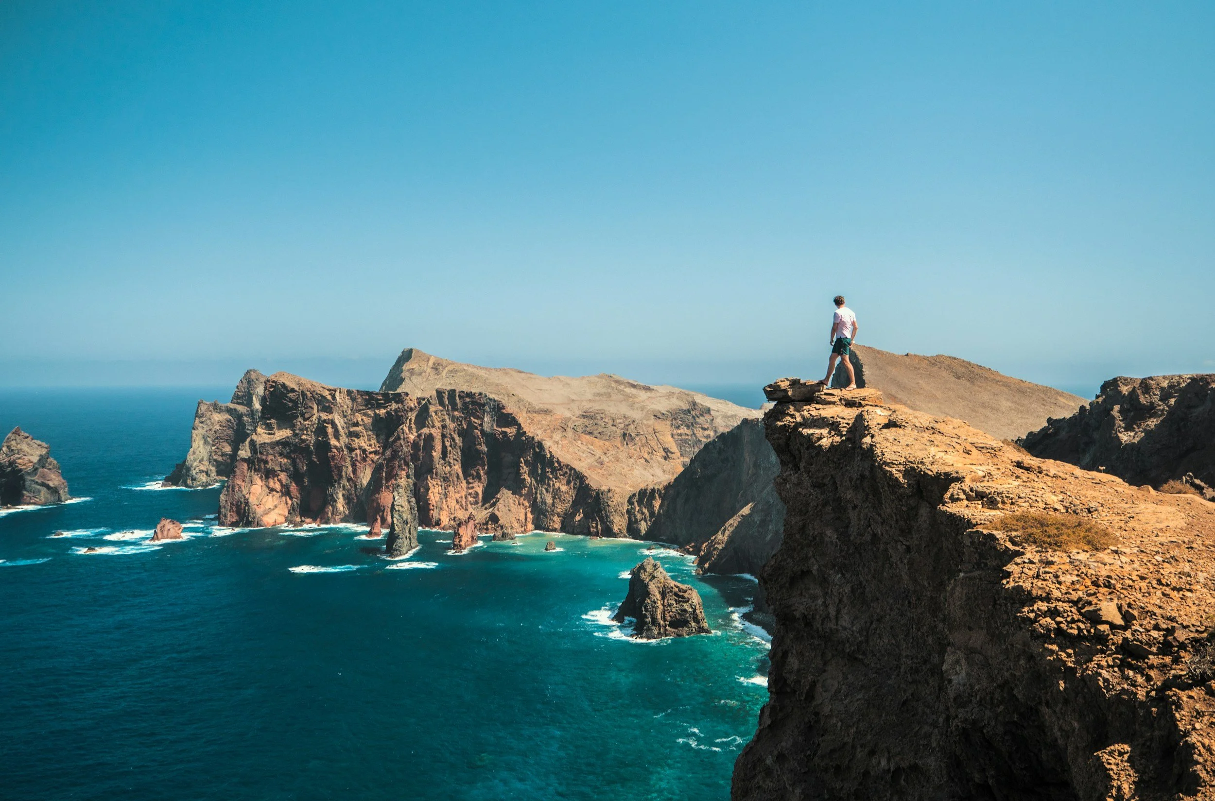 A person standing on a rocky cliff overlooking the ocean with dramatic rock formations and cliffs in the background under a clear blue sky.