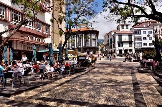 Tables under awnings on the pedestrian square outside Apolo Café, with Funchal Cathedral behind them