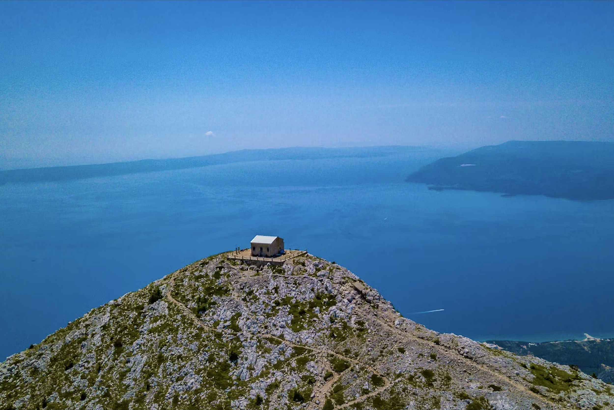 A small house on a hill overlooking a large body of water with distant landforms on the horizon under a clear blue sky.