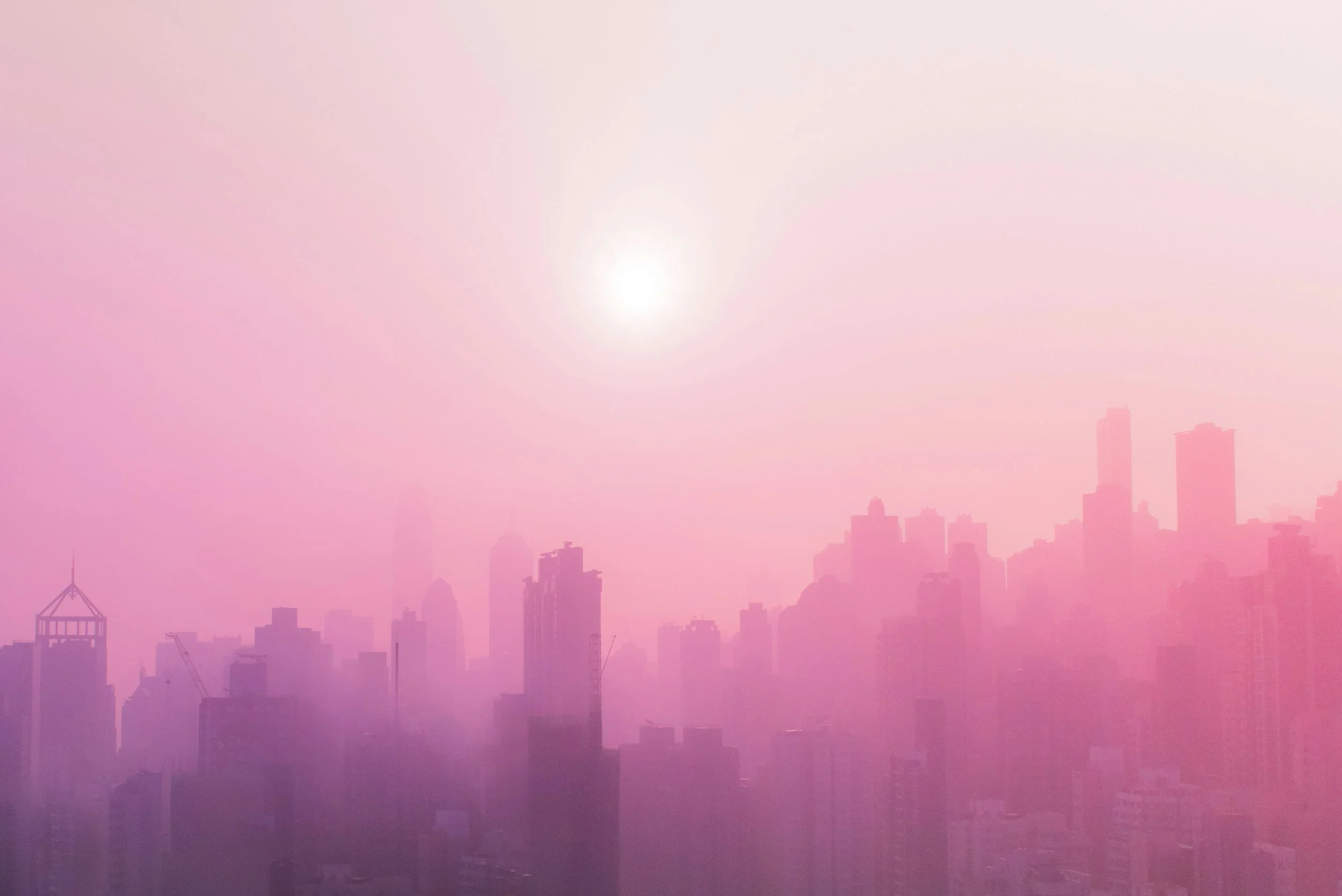 City skyline with tall buildings shrouded in pinkish haze and a bright sun in the sky.