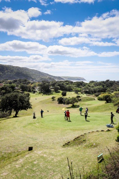 Golfers on a green overlooking a scenic landscape with hills and the ocean in the background on a sunny day.