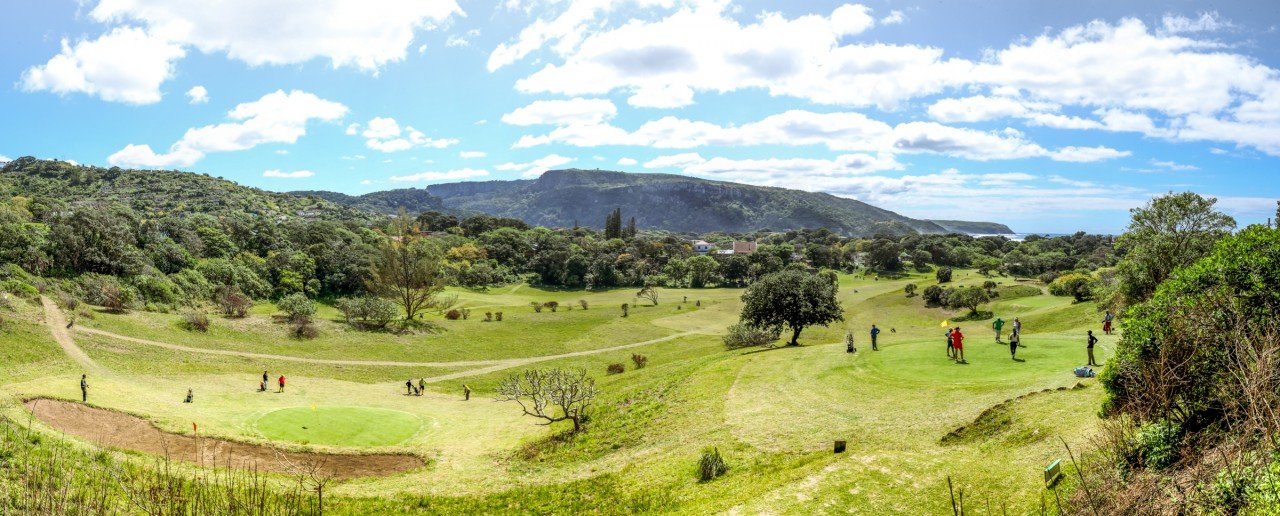 A scenic view of a golf course with golfers playing, surrounded by lush greenery and hills under a partly cloudy sky.