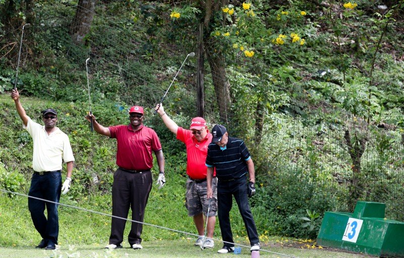 Four men on a golf course posing with clubs raised, wearing casual attire, standing near a green tee marker labeled with the number 3, surrounded by trees and greenery.