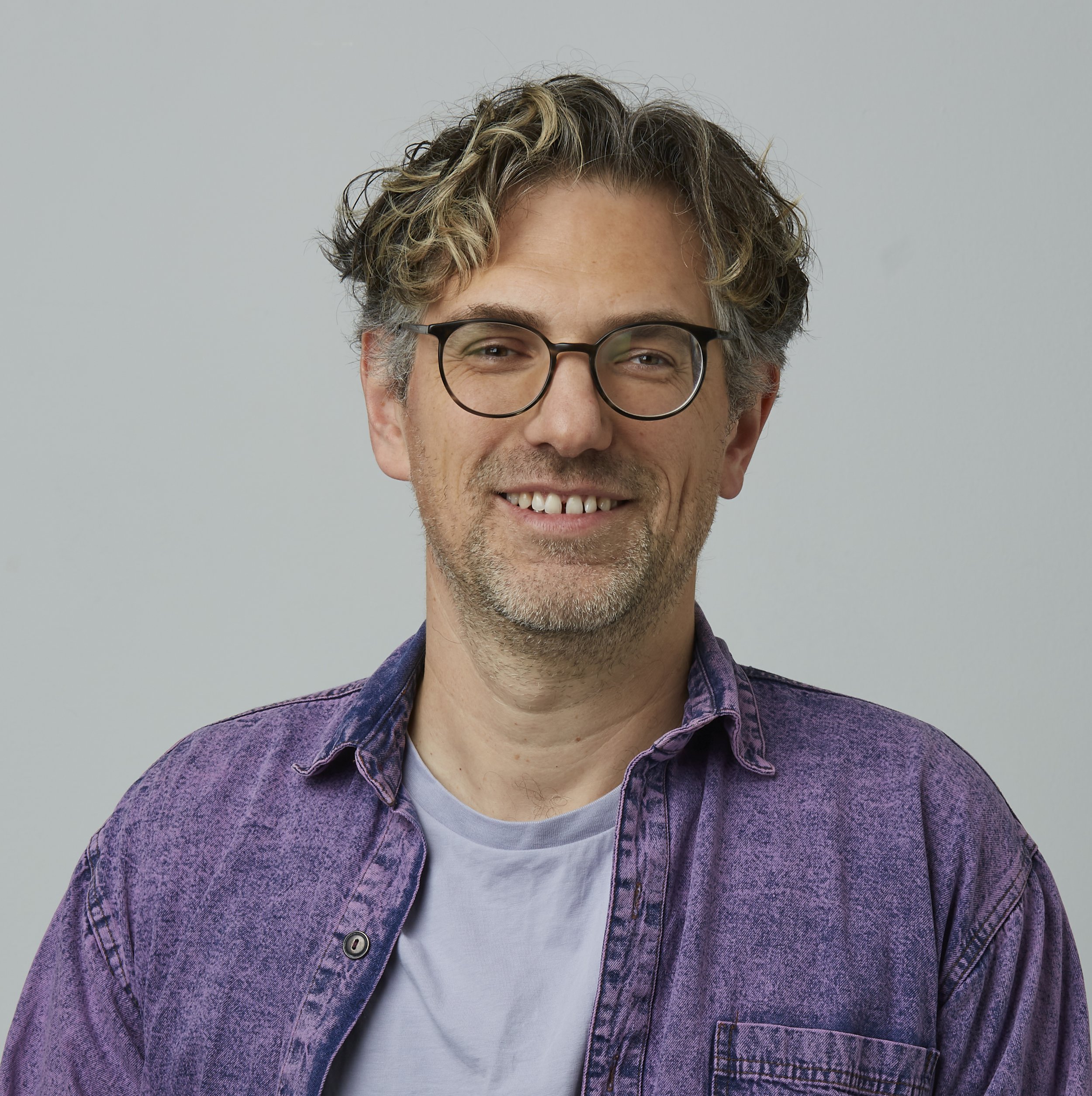 Dr. Jonas Westphal, Coach und Organisatonsentwickler schaut lächelnd in die Kamera. Er trägt eine dunkle Brille und ein offened lilafarbenes Jeanshemd.