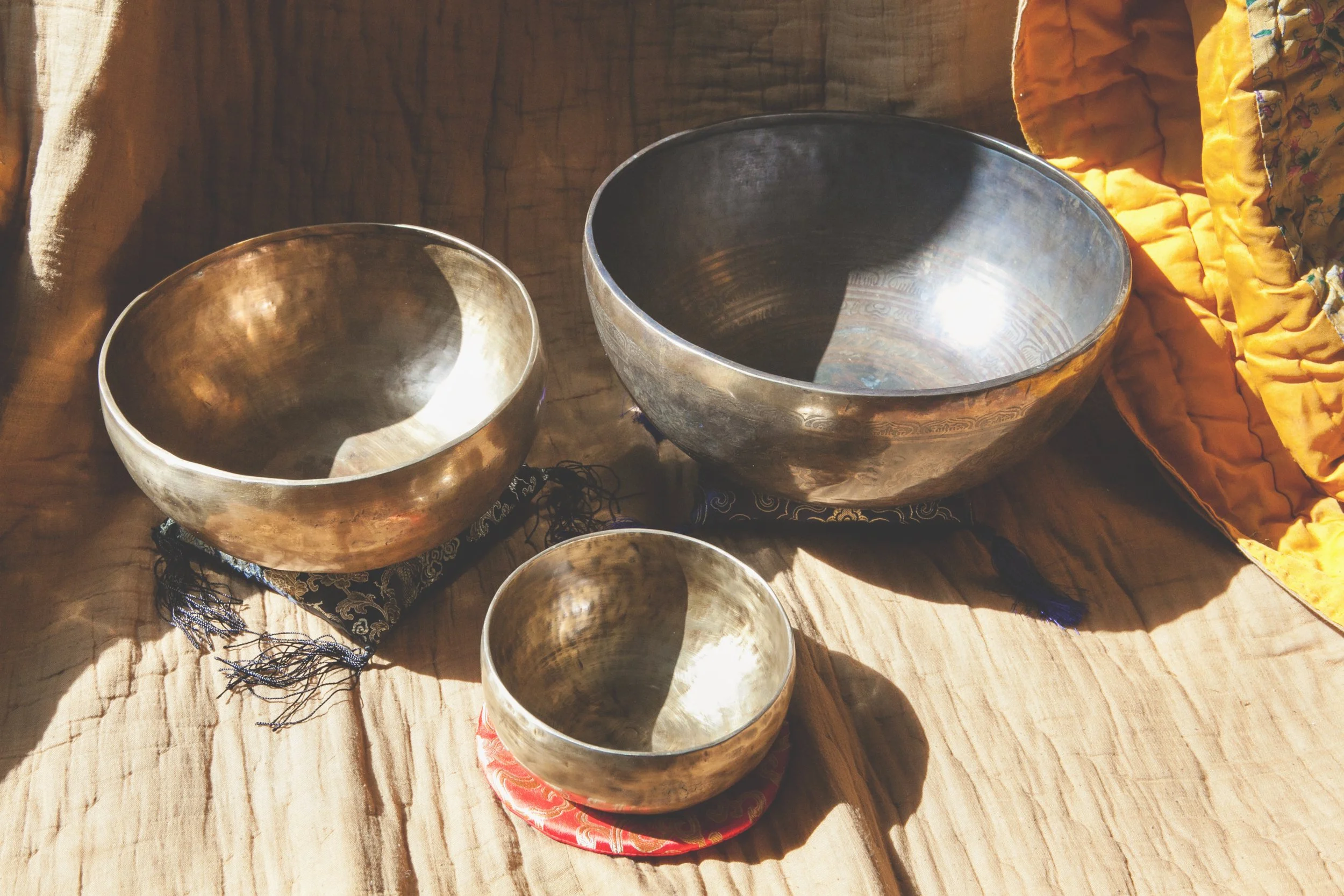 Three Himalayan singing bowls on a wooden surface, with sunlight reflecting off their metal surfaces.